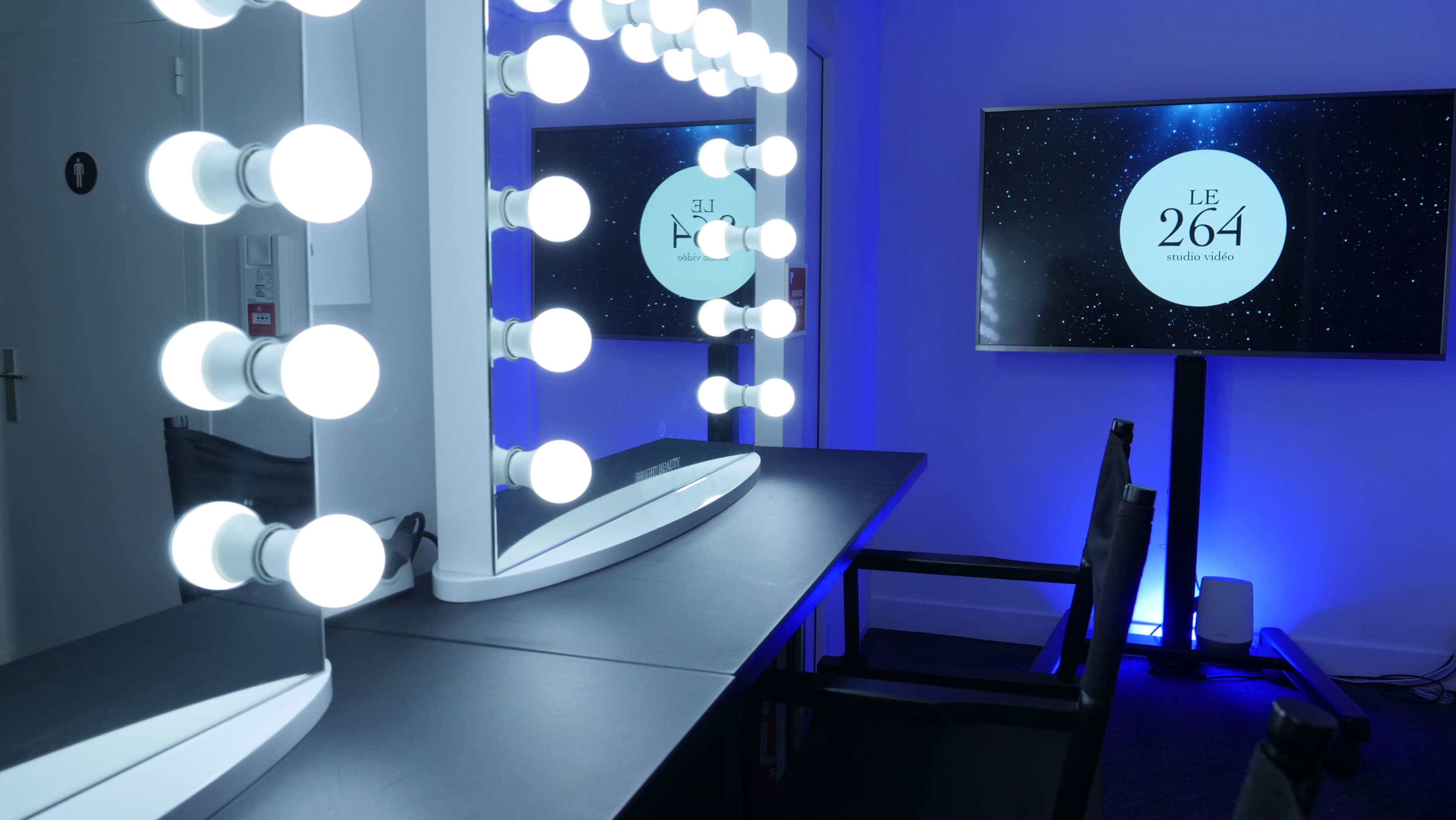 The image shows a well-lit makeup vanity with large circular lights and a nearby screen displaying the text "LE 264 studio vidéo."
