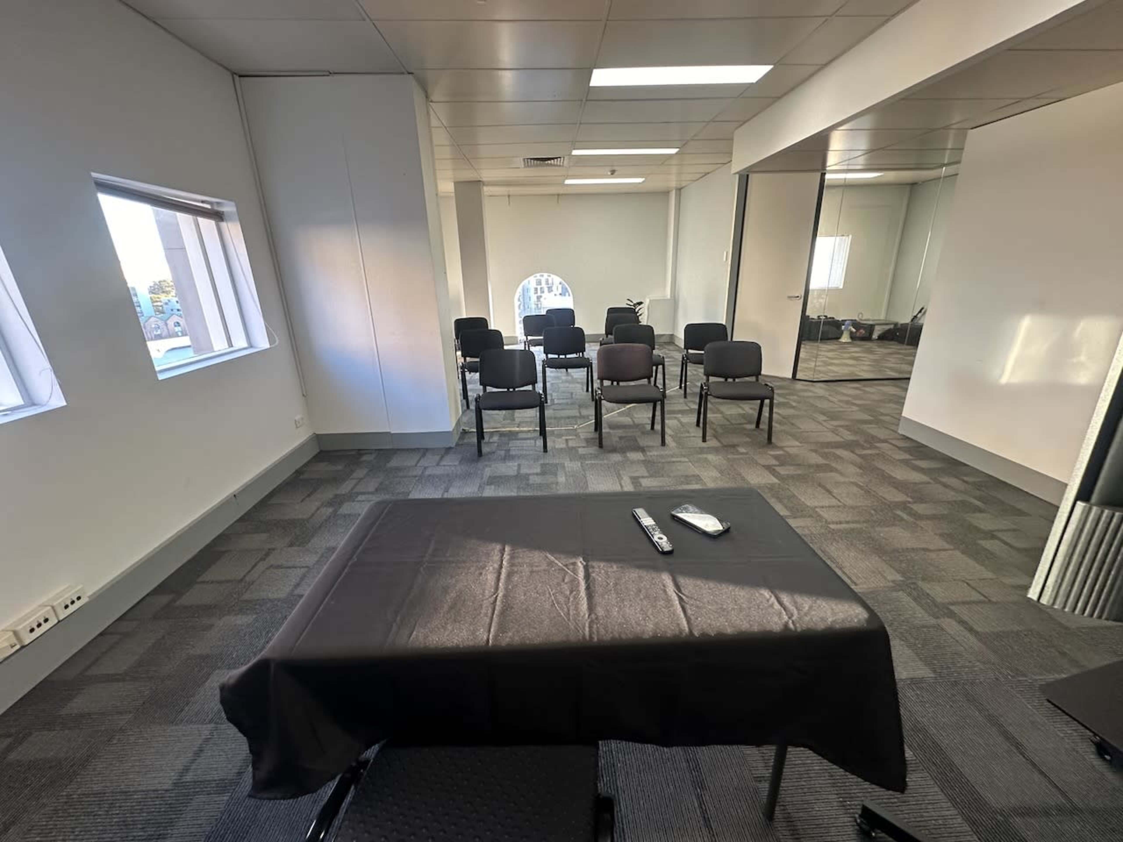 A conference room features a black table at the center, with a notepad and pen, surrounded by several chairs arranged in rows against the wall.