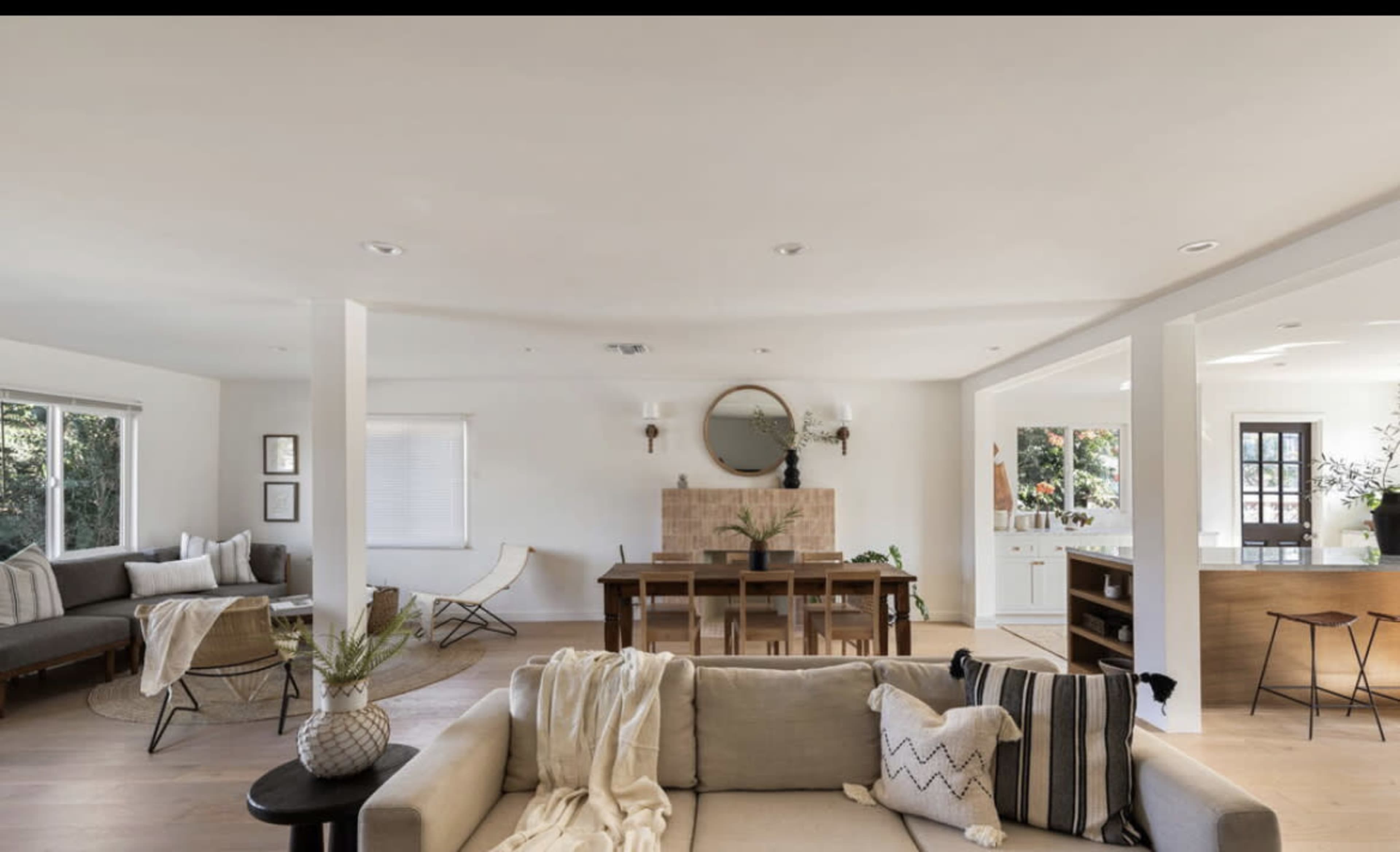 The image shows a modern, open-concept living and dining area with a neutral color palette, featuring a sofa, dining table, and large windows.
