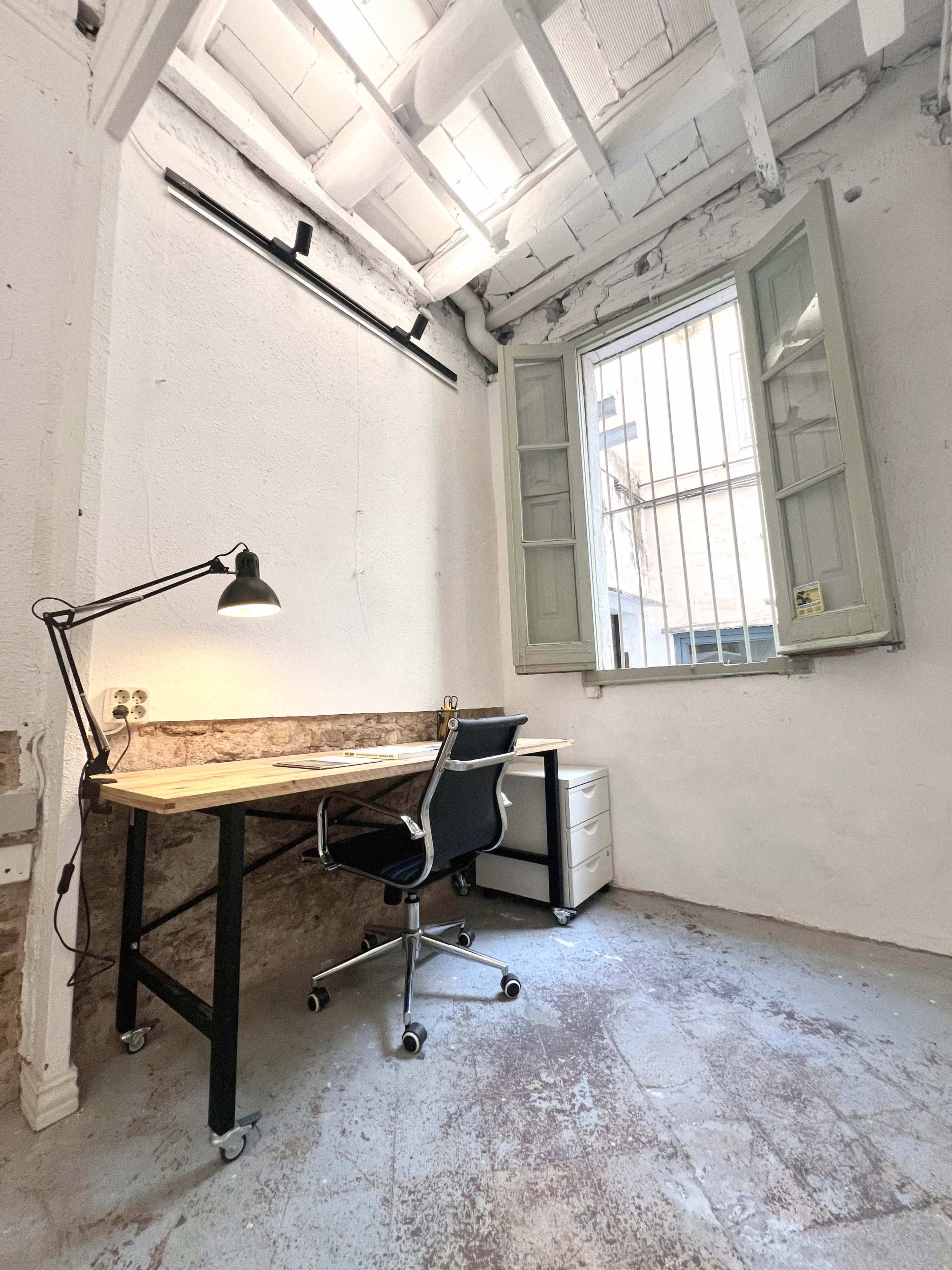 The image shows a minimalistic office space with a wooden desk, a black chair, a lamp, and a small filing cabinet under a window with open shutters.