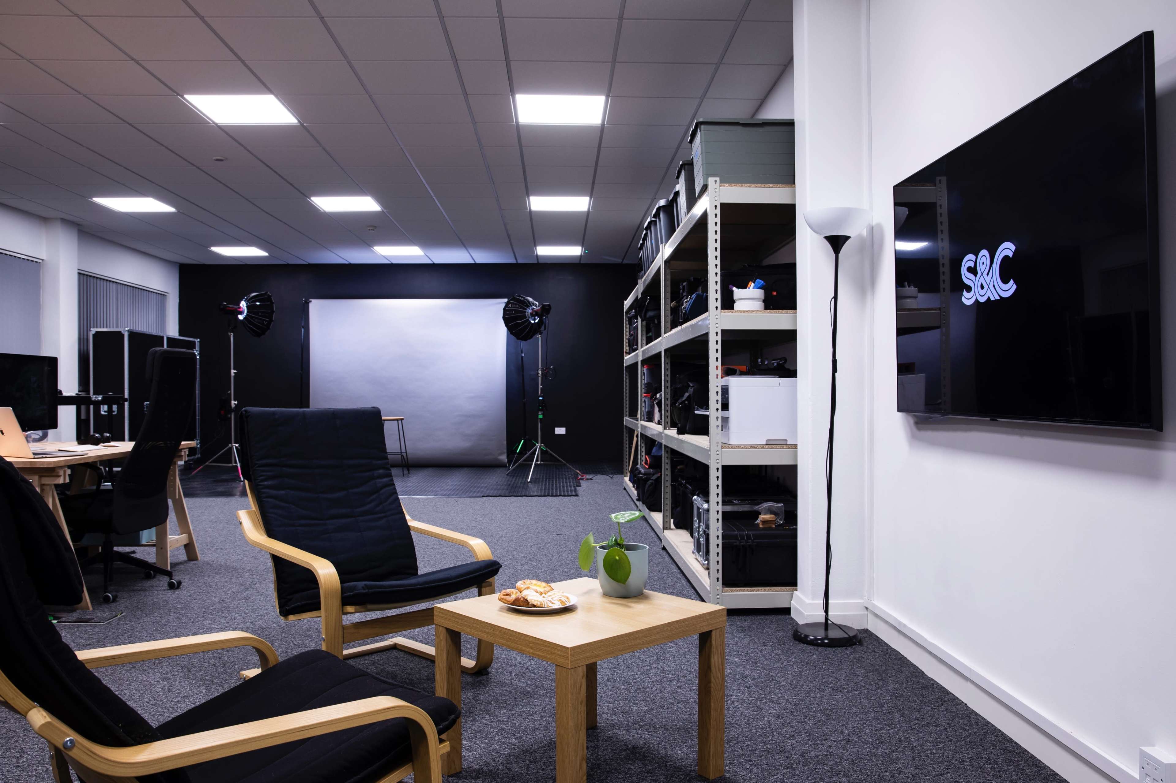 A modern office space with black chairs, a small table holding snacks, and equipment shelves, alongside a backdrop and studio lights set up for photography.