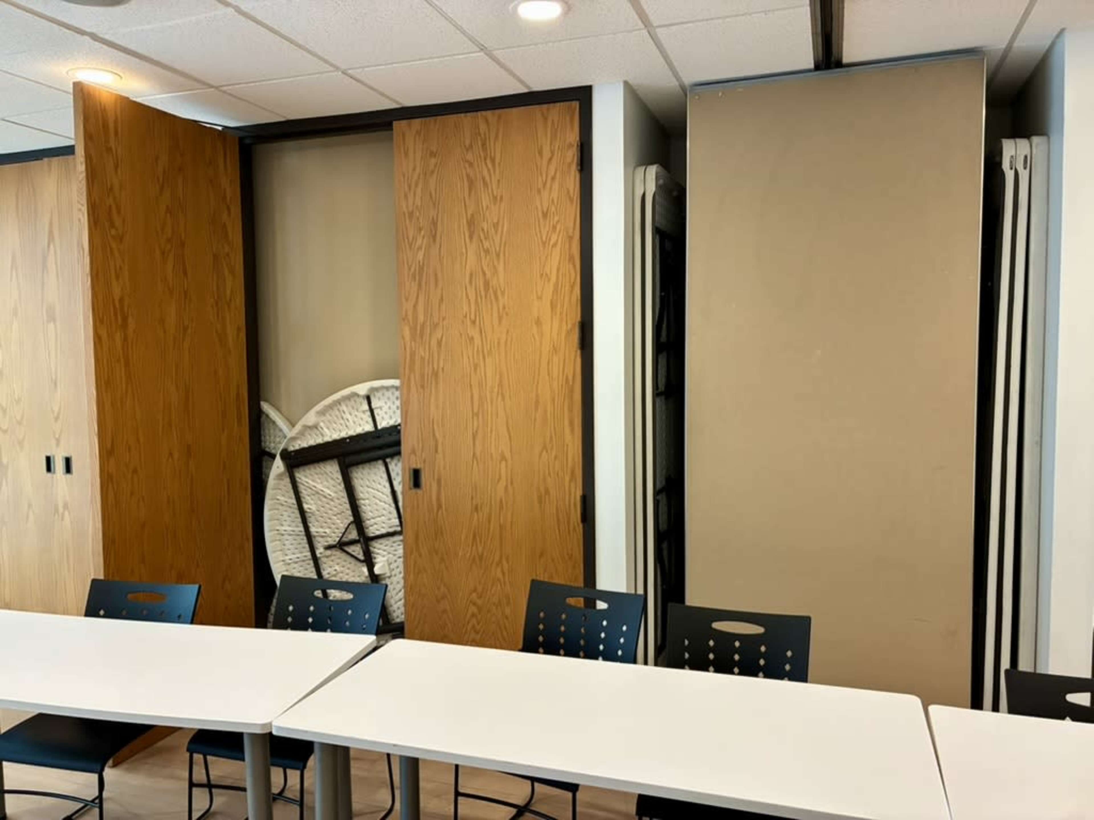 A room featuring a long white table with black chairs, flanked by wooden foldable doors and what appears to be a stacked folding table stored against the wall.
