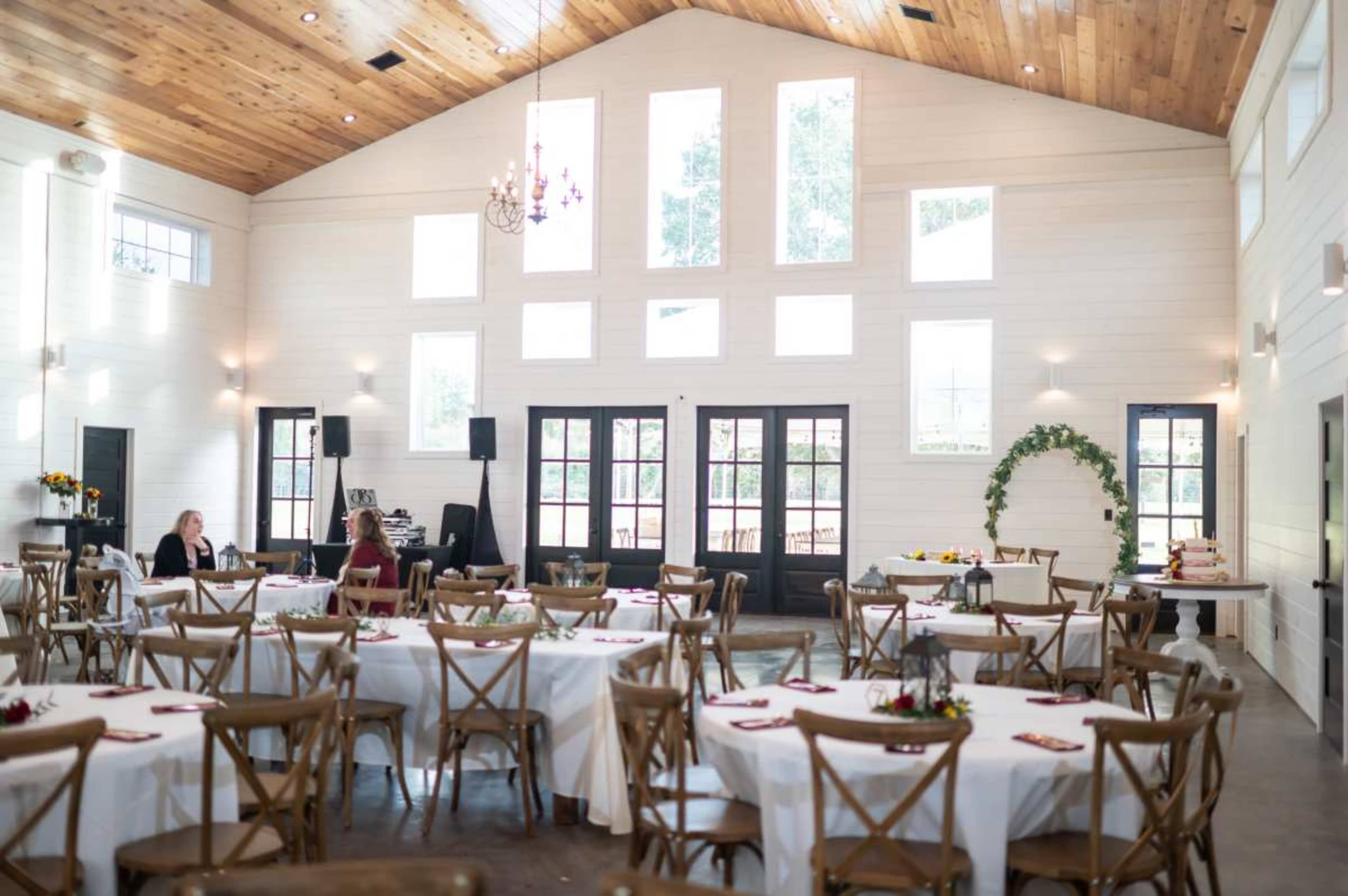 The interior of a banquet hall features circular tables with white tablecloths, surrounded by wooden chairs, and large windows allow natural light to fill the space.