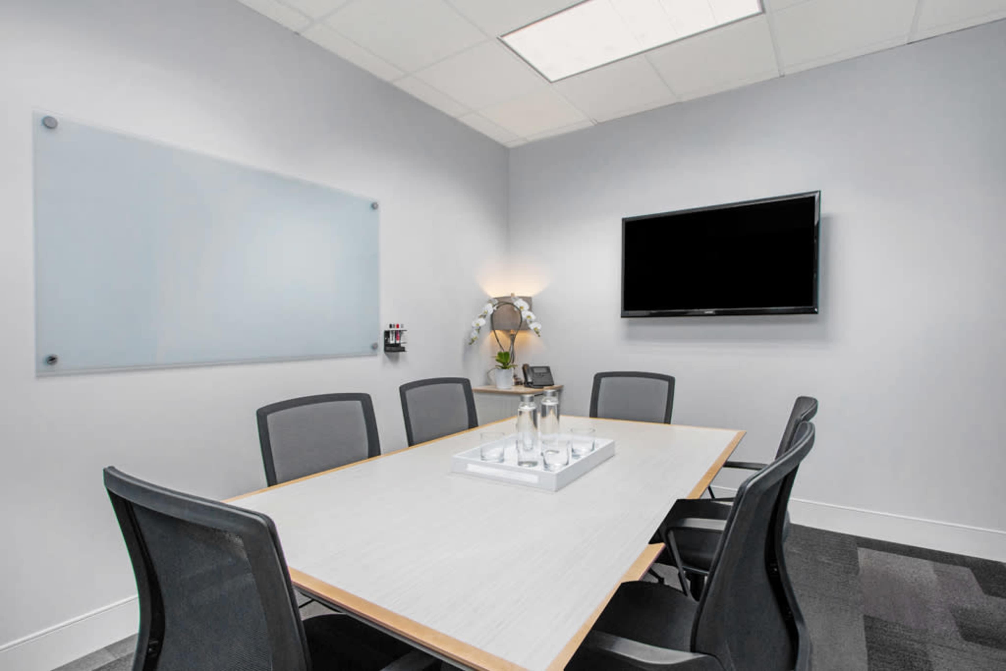 A modern conference room features a large table surrounded by ergonomic chairs, a wall-mounted TV, and a whiteboard.