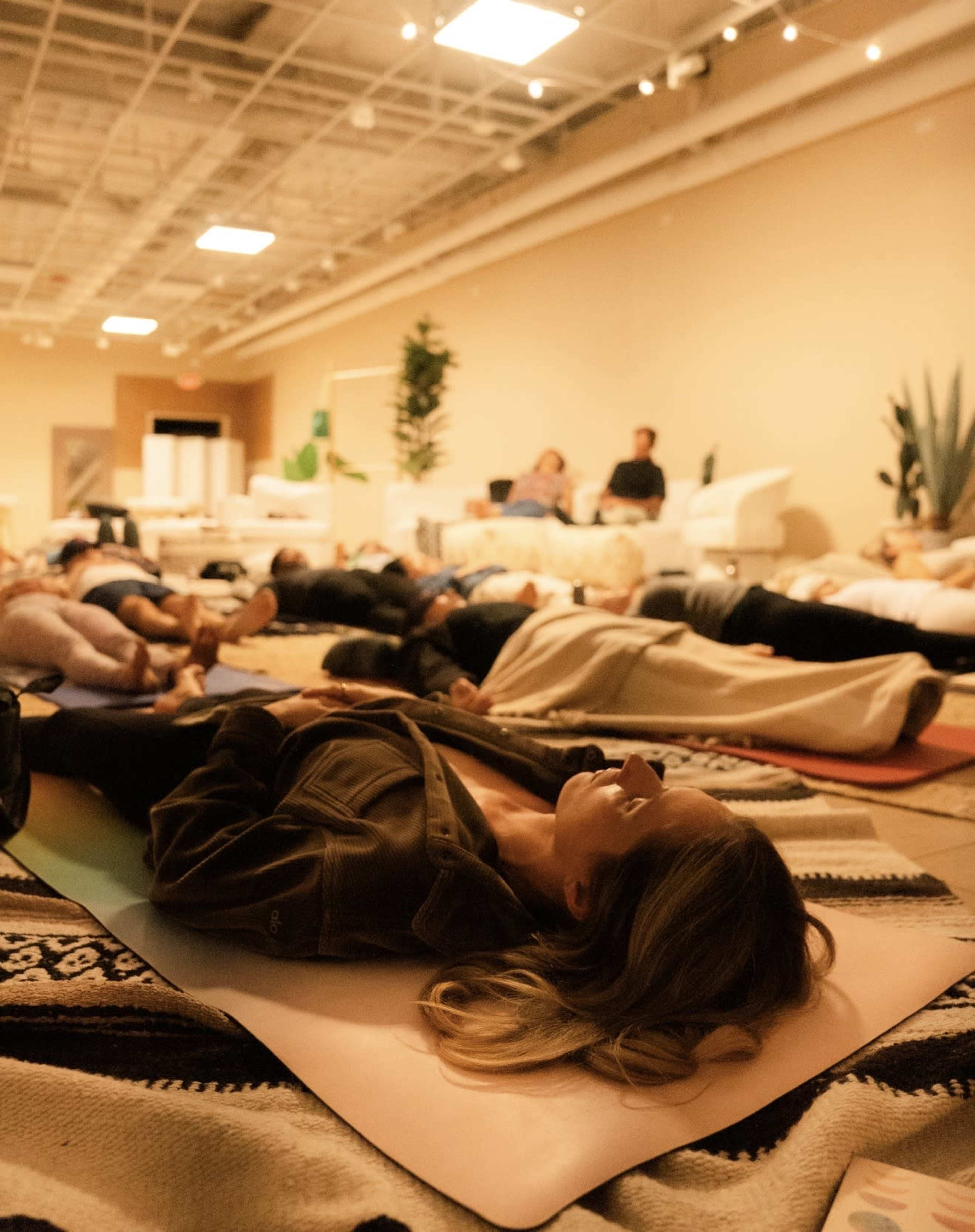 A group of people is lying on yoga mats in a spacious room, practicing relaxation or meditation.