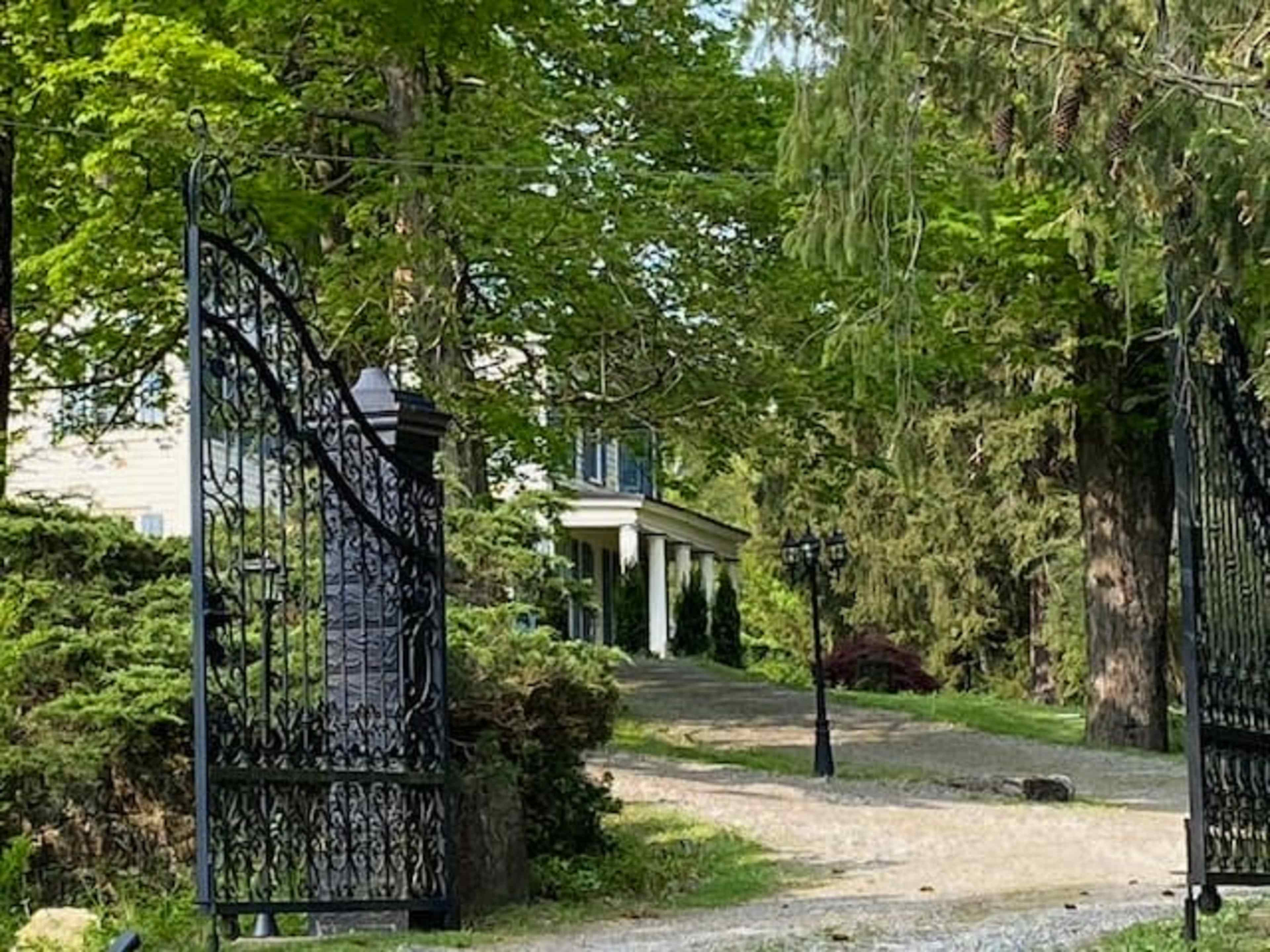 An ornate black iron gate opens onto a gravel path leading to a house surrounded by lush greenery.