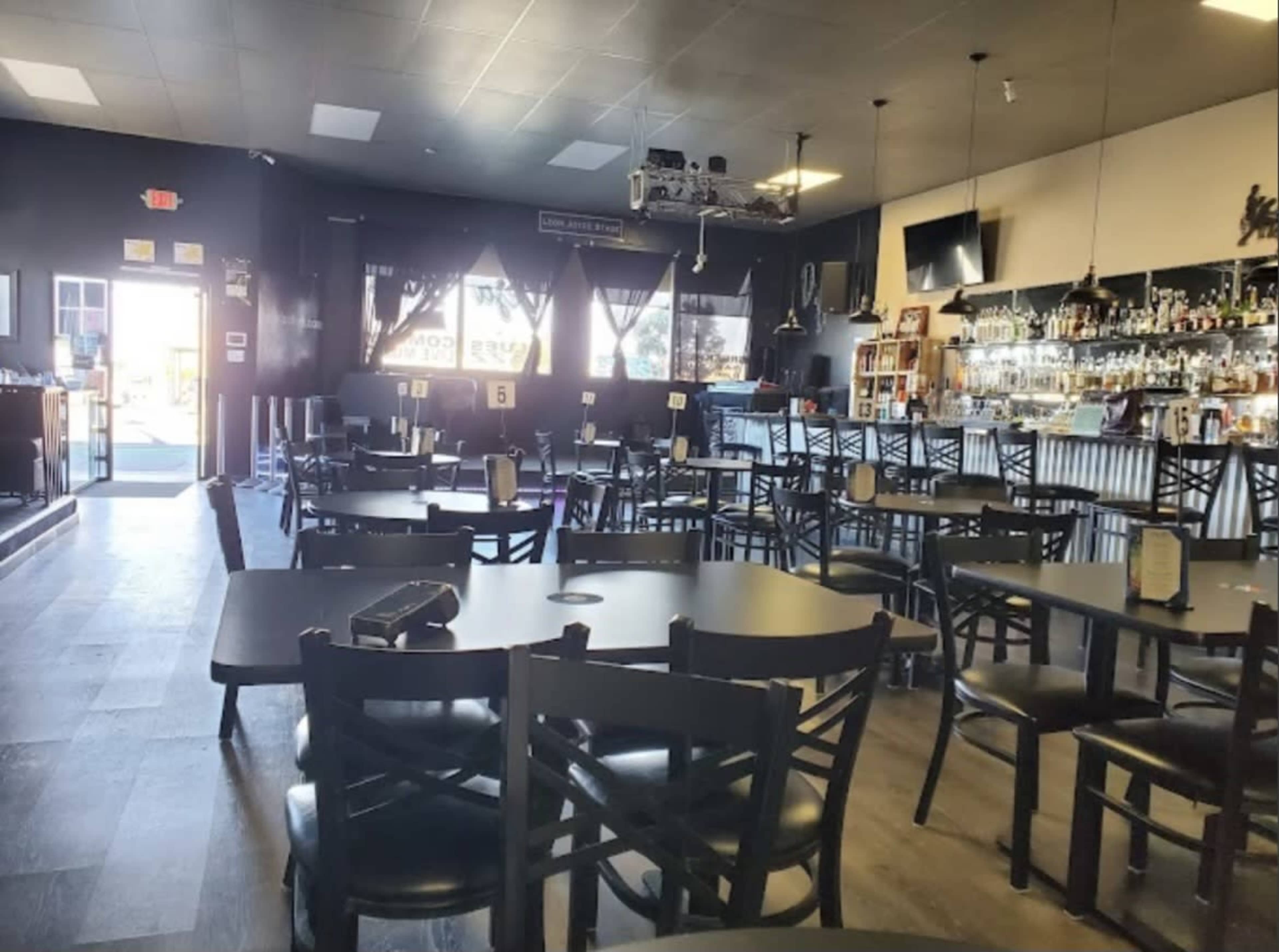 The image shows a spacious restaurant interior featuring numerous black tables and chairs, a bar area with shelves of bottles, and large windows at the entrance.