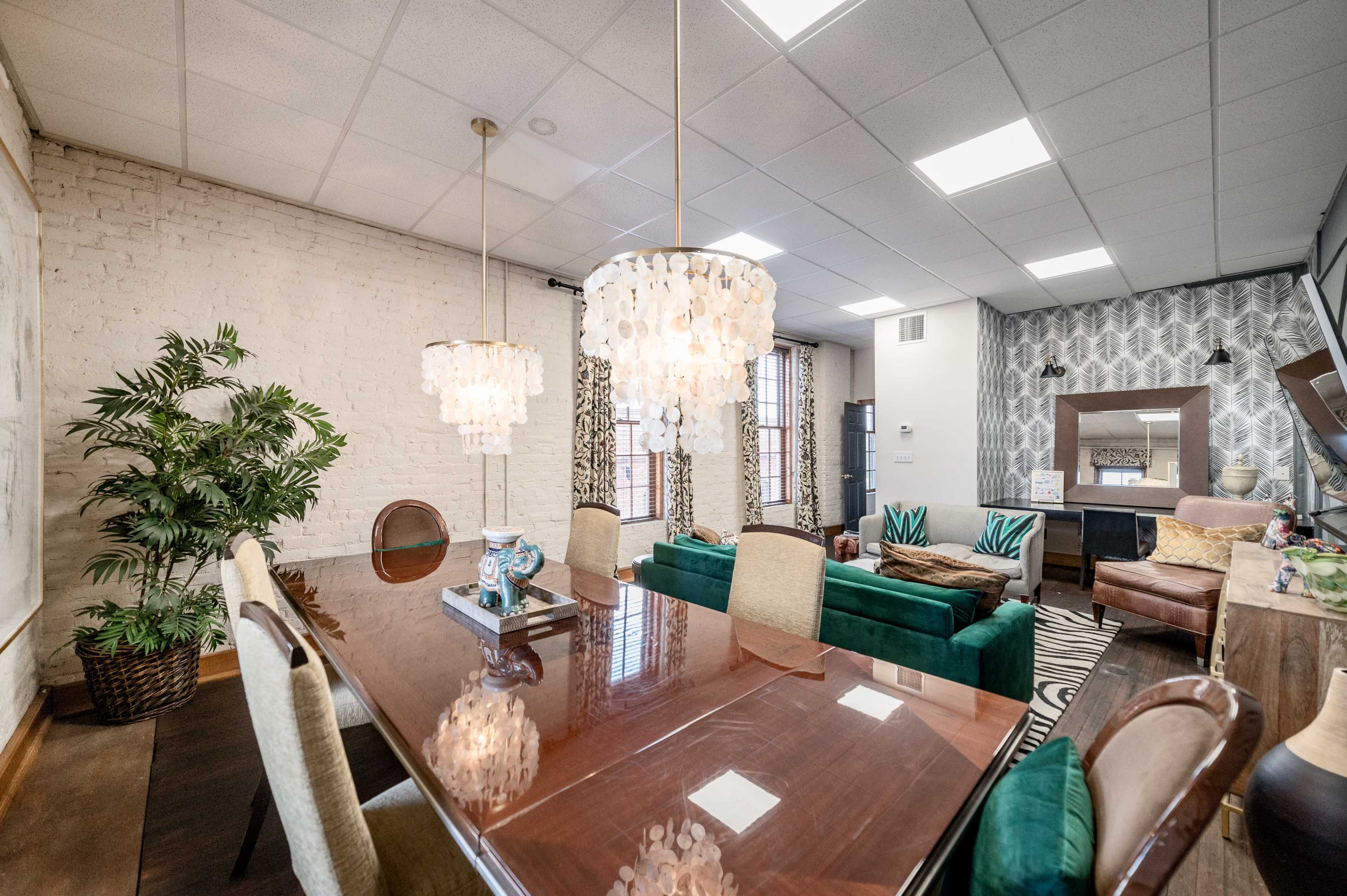 A modern dining area features a long wooden table with upholstered chairs, surrounded by decorative lighting and a potted plant.