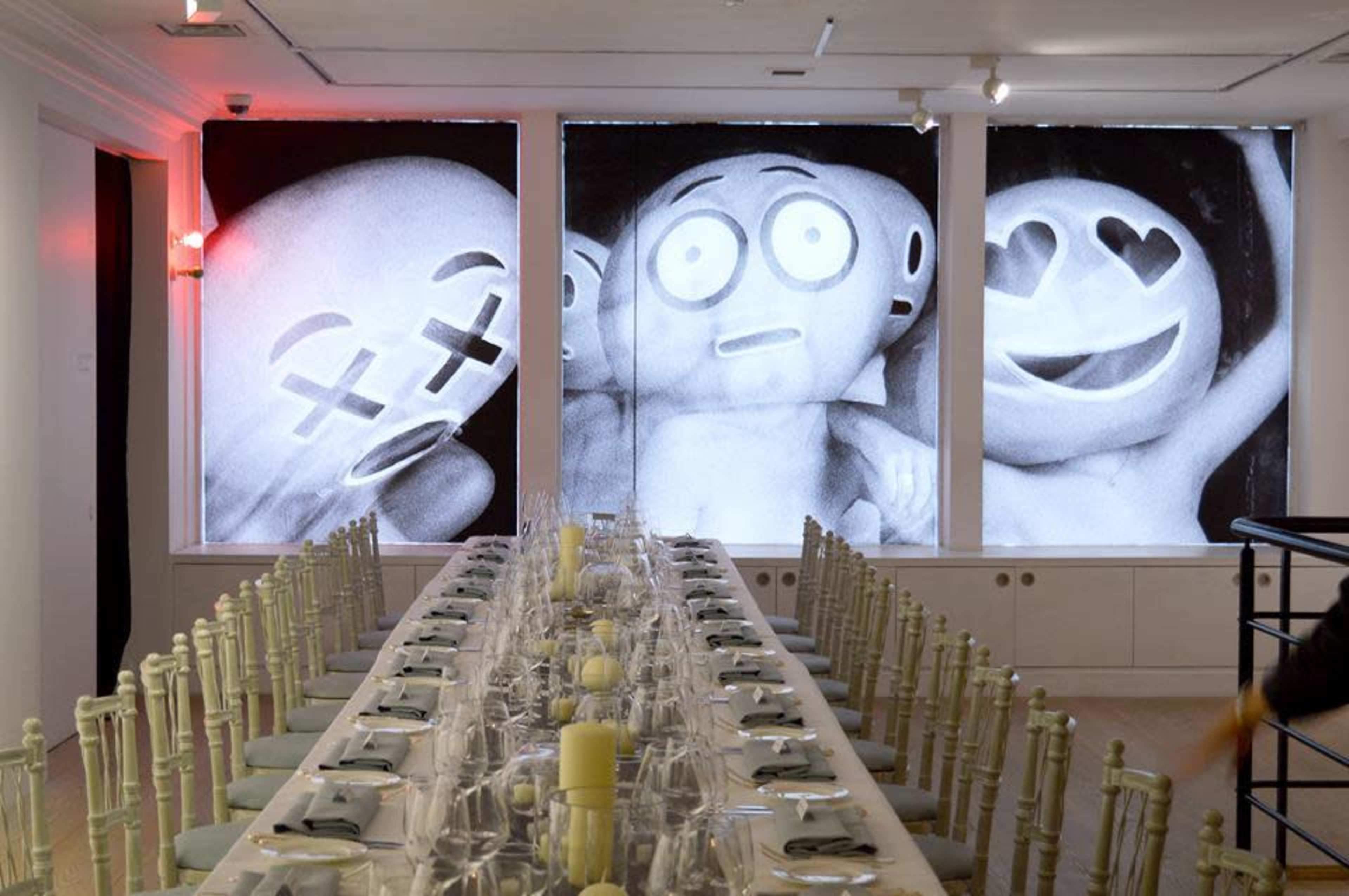 A dining area with a long table set for a meal, featuring three large black-and-white emoji-like figures displayed on the windows.