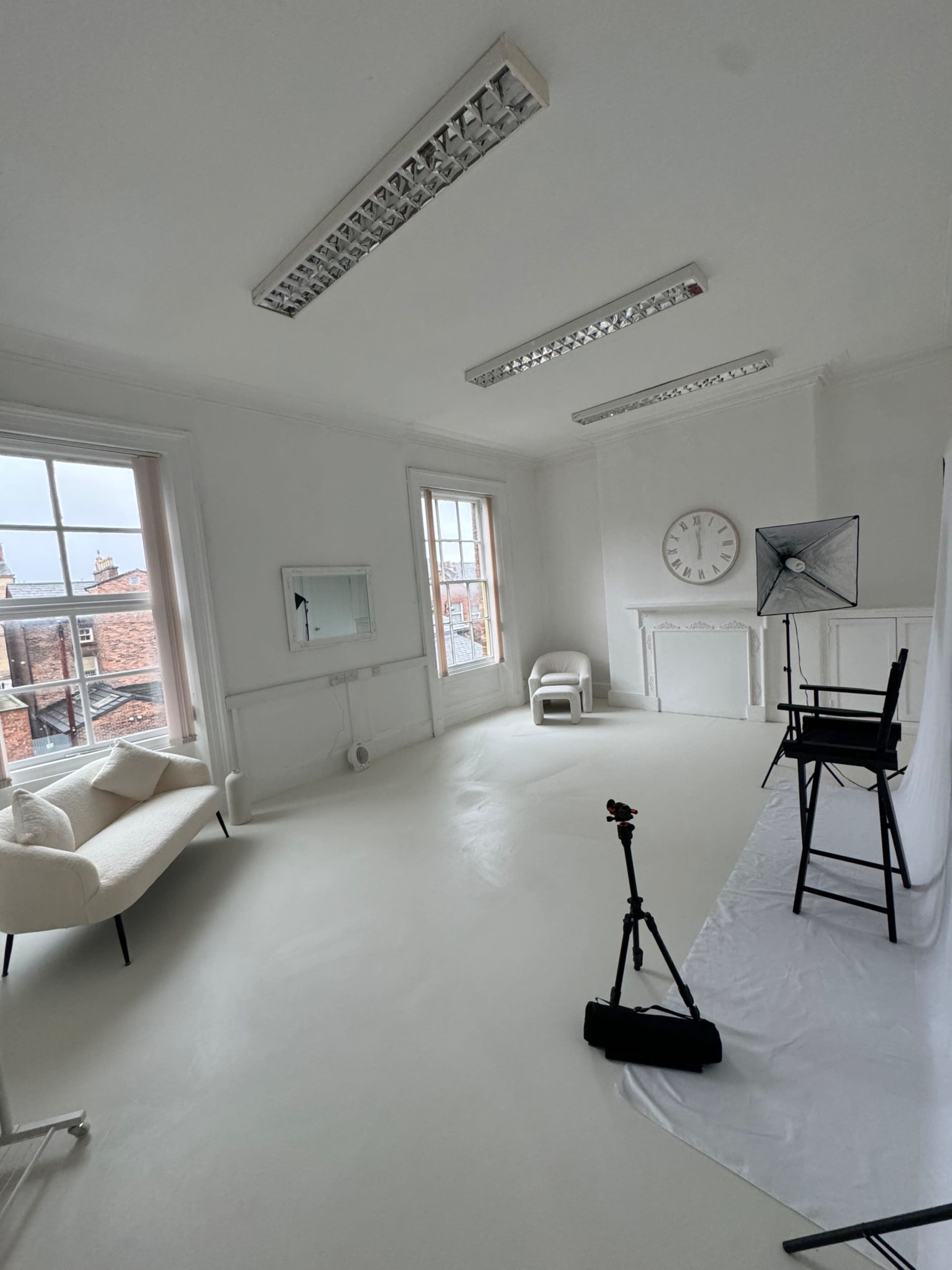 The image shows a bright, minimalist studio space with white walls and flooring, featuring a chair, a mirror, a clock, and a photography setup in one corner.