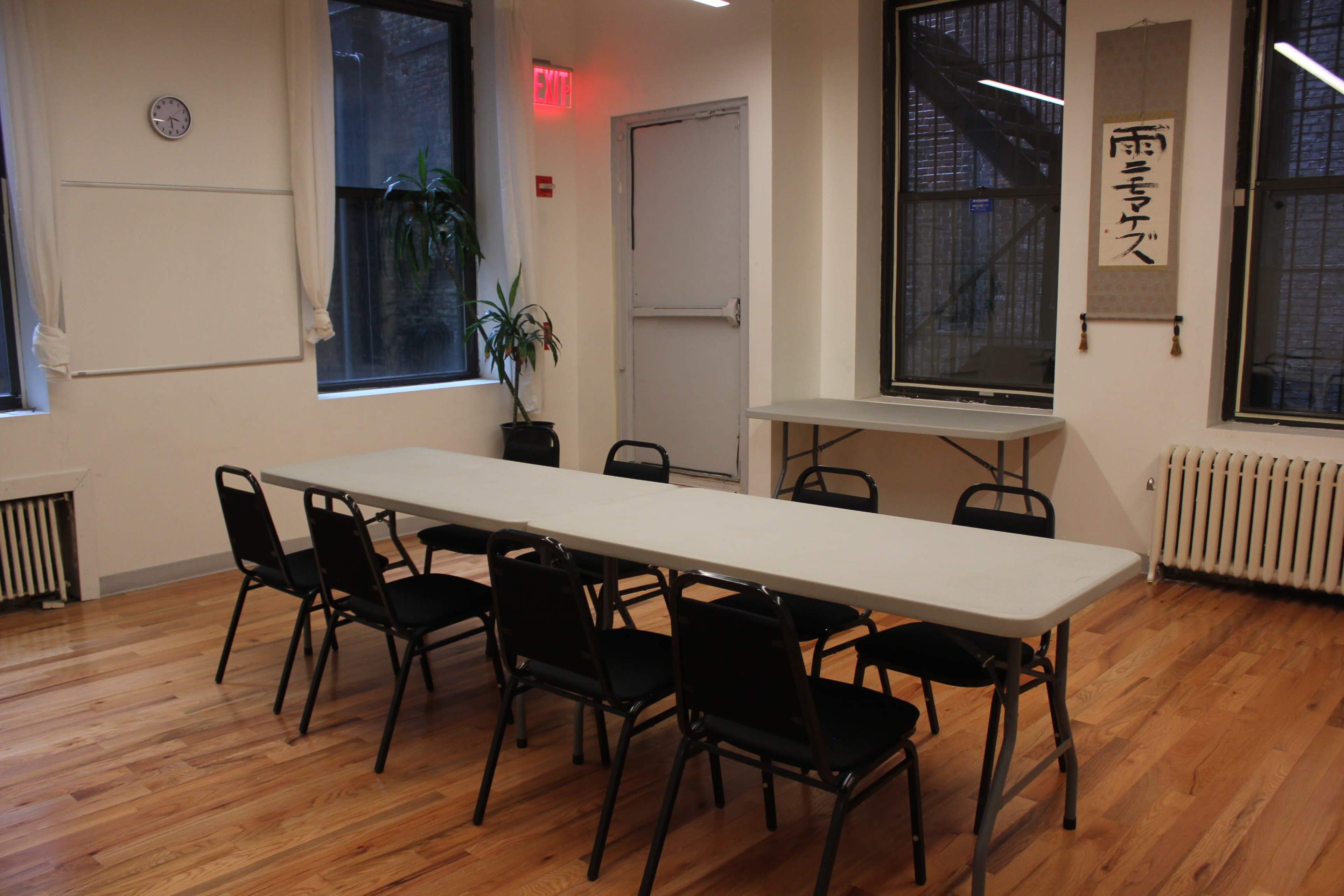 A large, empty table surrounded by black chairs is set up in a well-lit room with wooden floors and windows.
