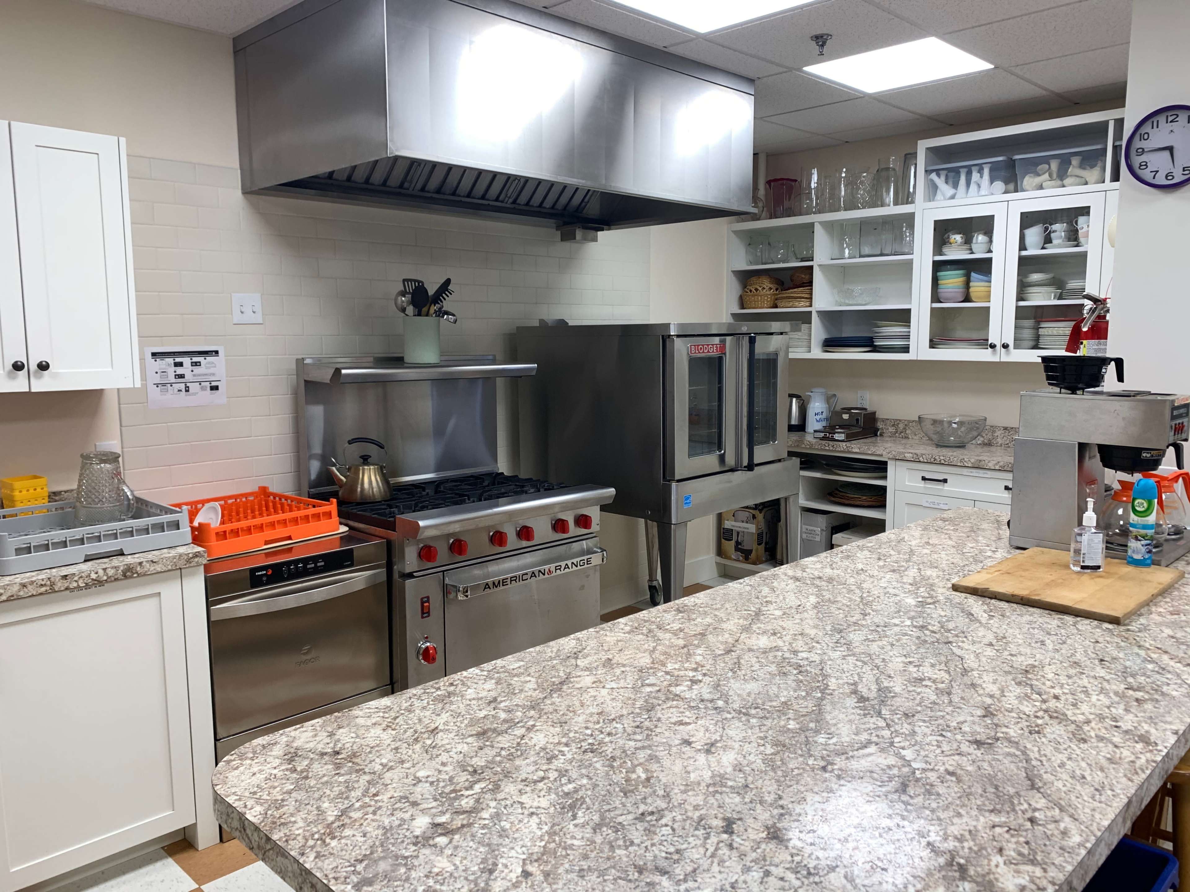 A commercial kitchen with a stainless steel ventilation hood, a gas range, a convection oven, and a granite countertop with various kitchen tools and equipment.