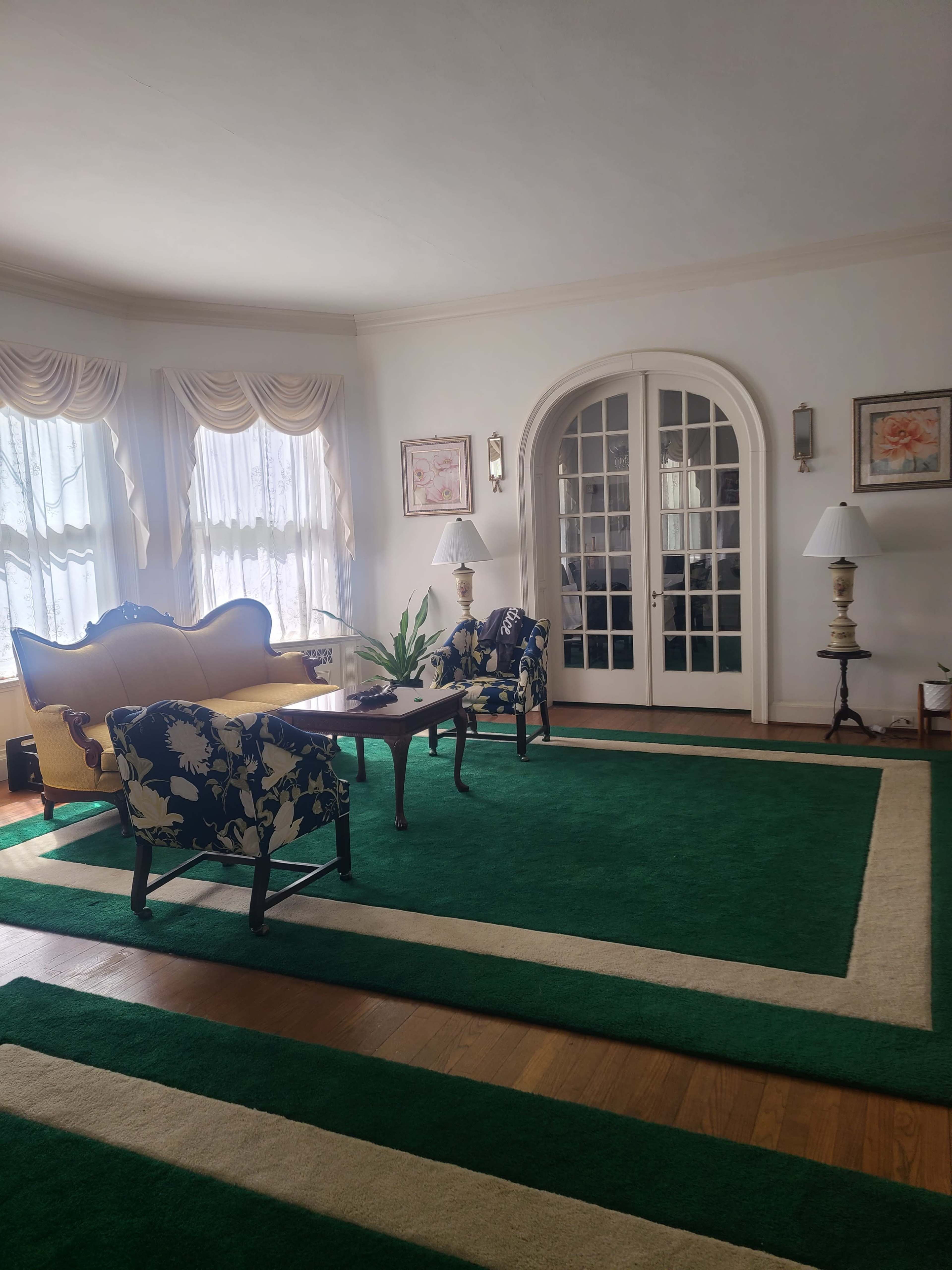 The image shows a living room with green carpet, two floral-patterned chairs, a small table, and a pair of lamps beside a set of glass-paneled French doors.