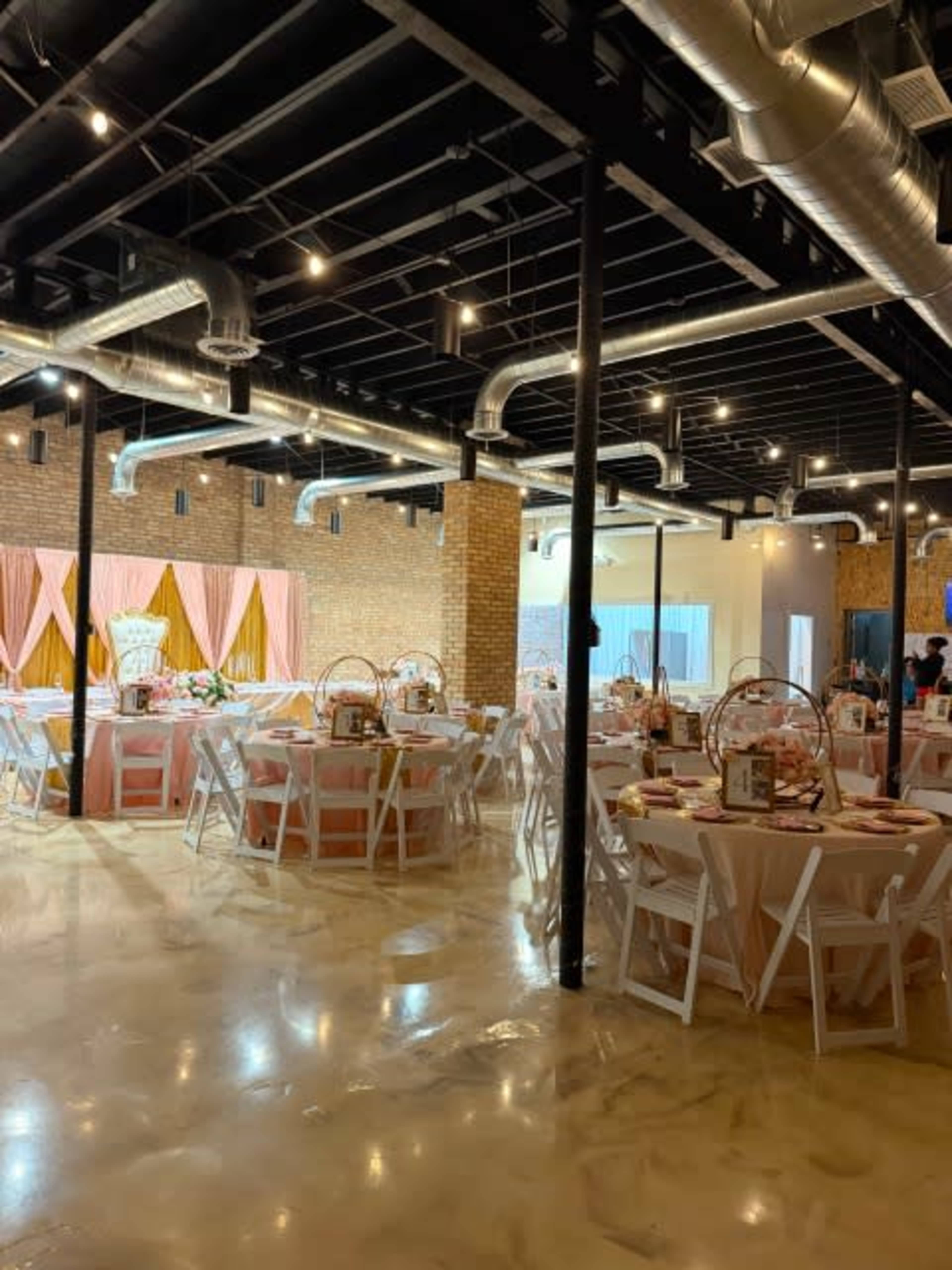 The image shows a spacious event venue set up for a celebration, featuring round tables adorned with pink tablecloths, chairs, and decorative elements, with a backdrop of draped fabric and exposed brick walls.