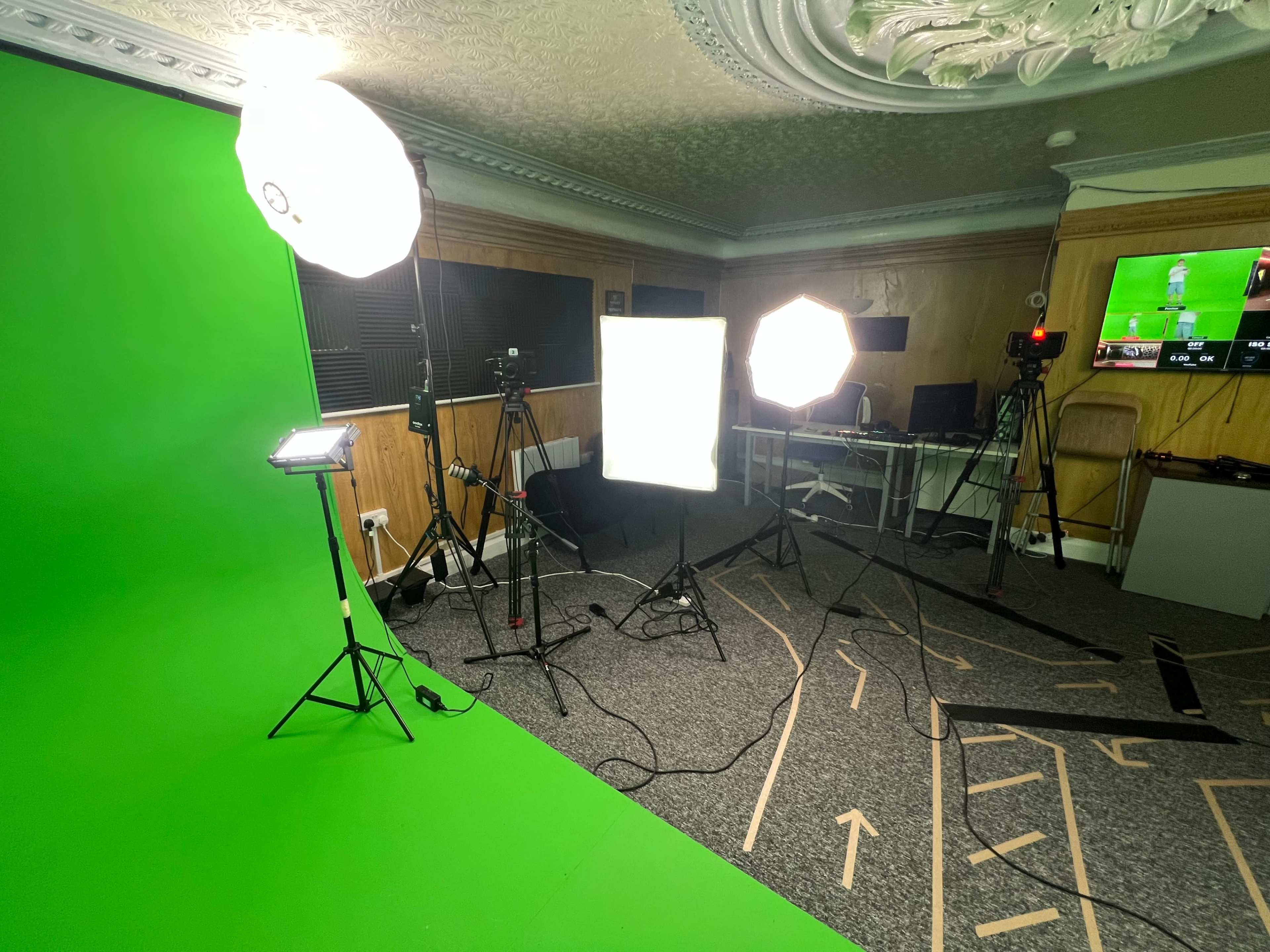 The image shows a room with a green screen setup, multiple lighting equipment, and cameras arranged for video production.