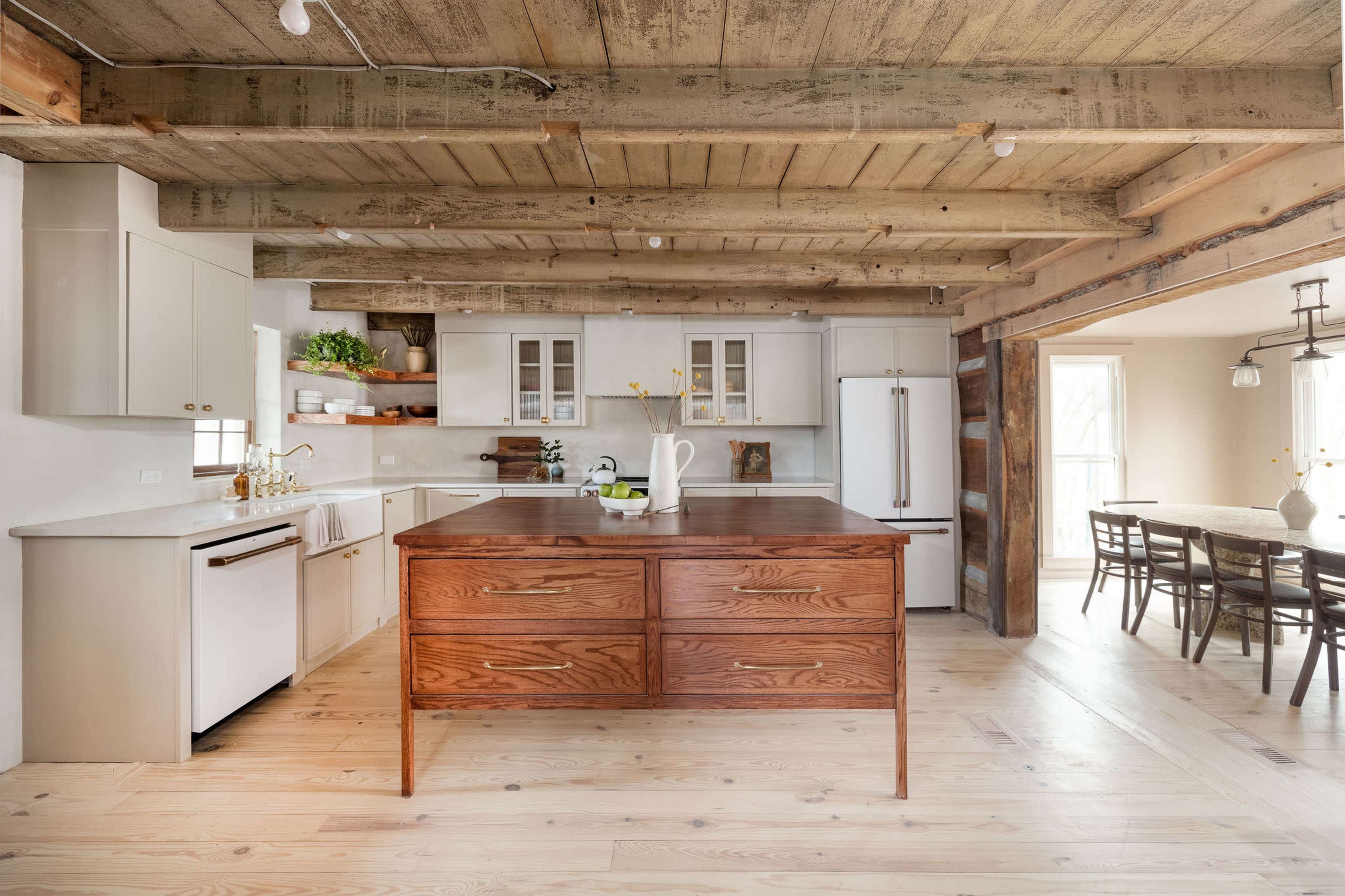 A modern kitchen with wooden beams, a large wooden island, and light-colored cabinetry, connected to a dining area with a wooden table and chairs.
