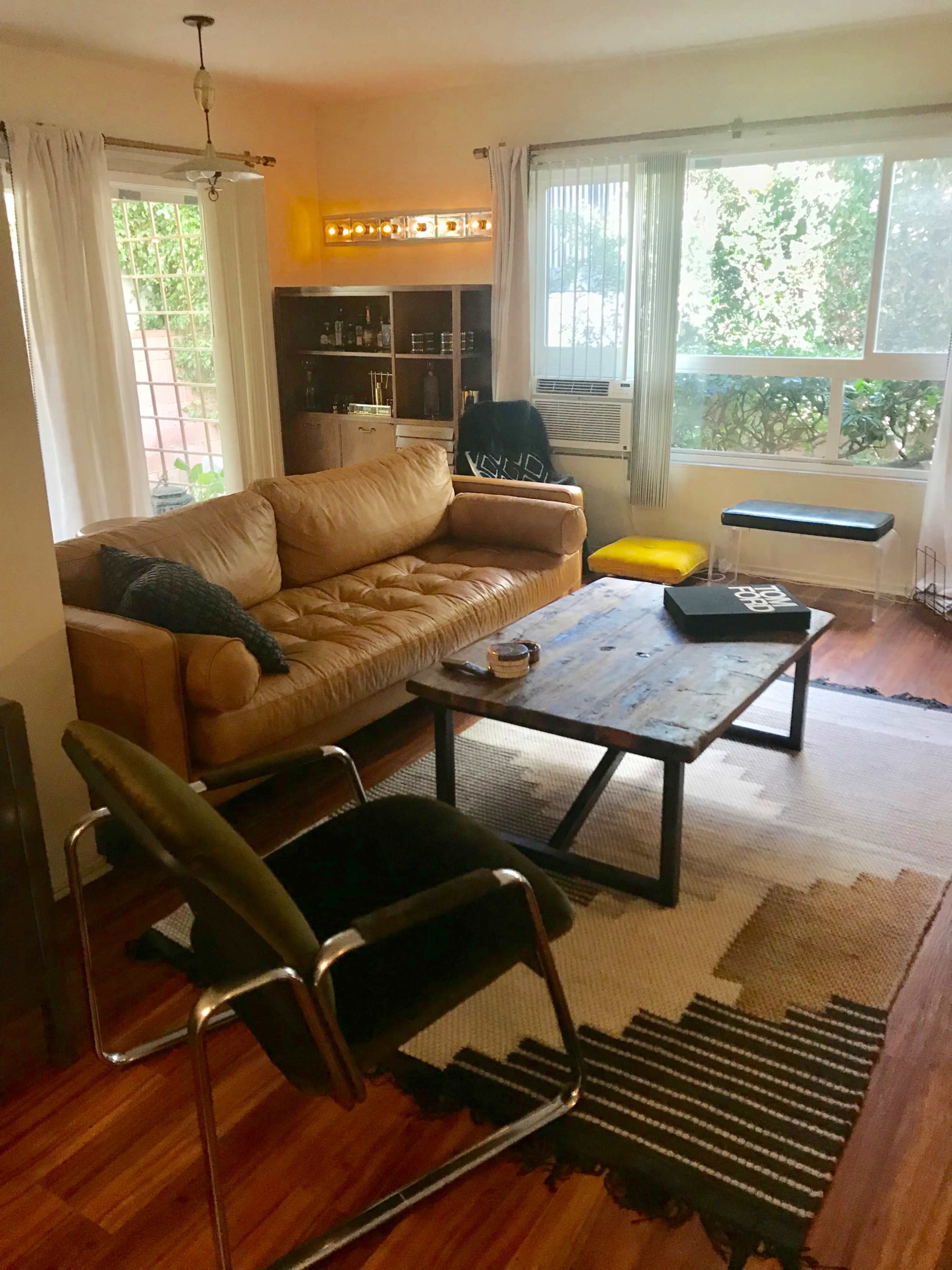 The image shows a living room with a large brown leather sofa, a wooden coffee table, and several pieces of furniture arranged around a patterned rug and natural light coming through large windows.