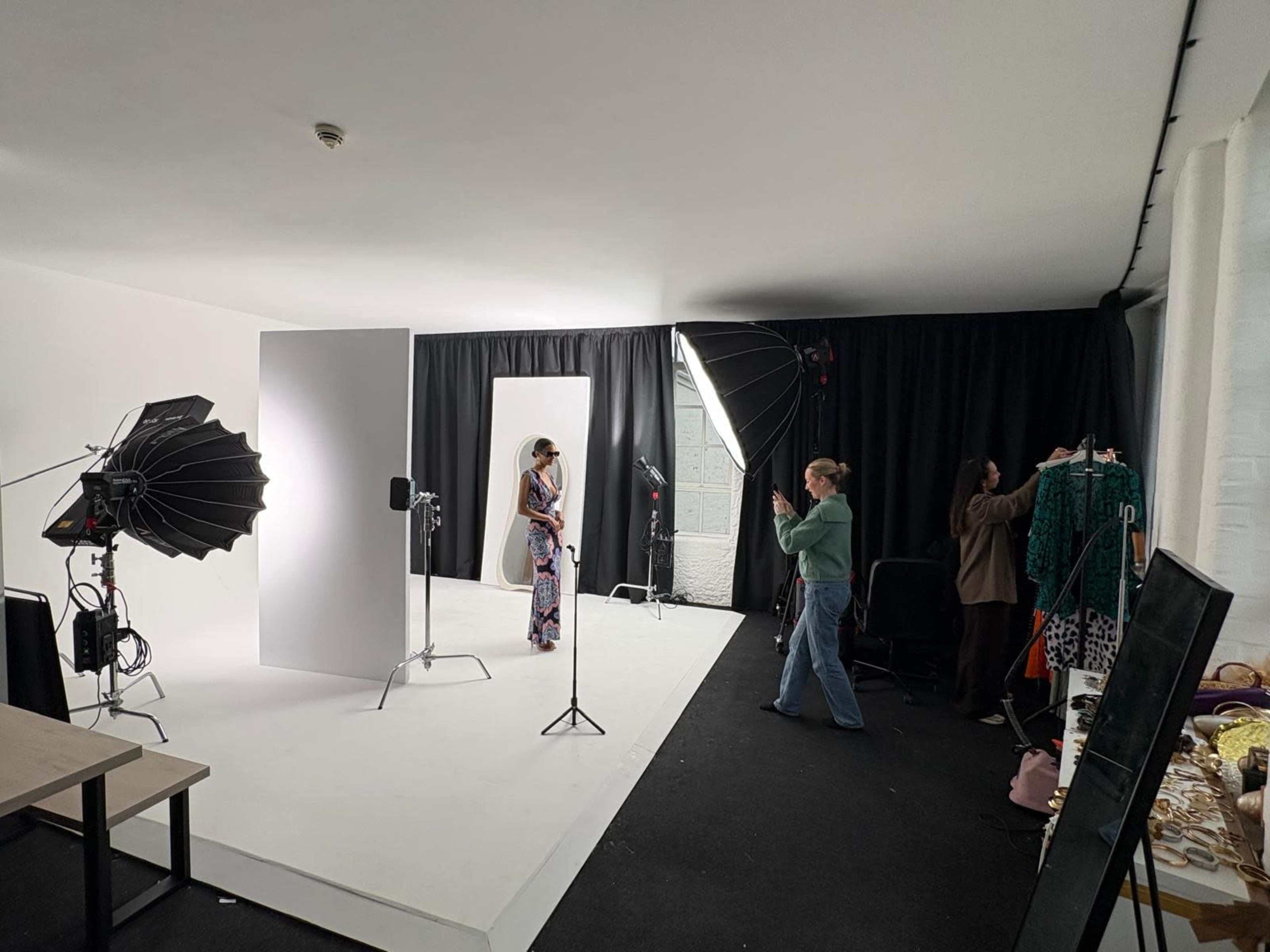 A model poses in front of a white backdrop while two stylists adjust outfits and lighting in a photography studio.