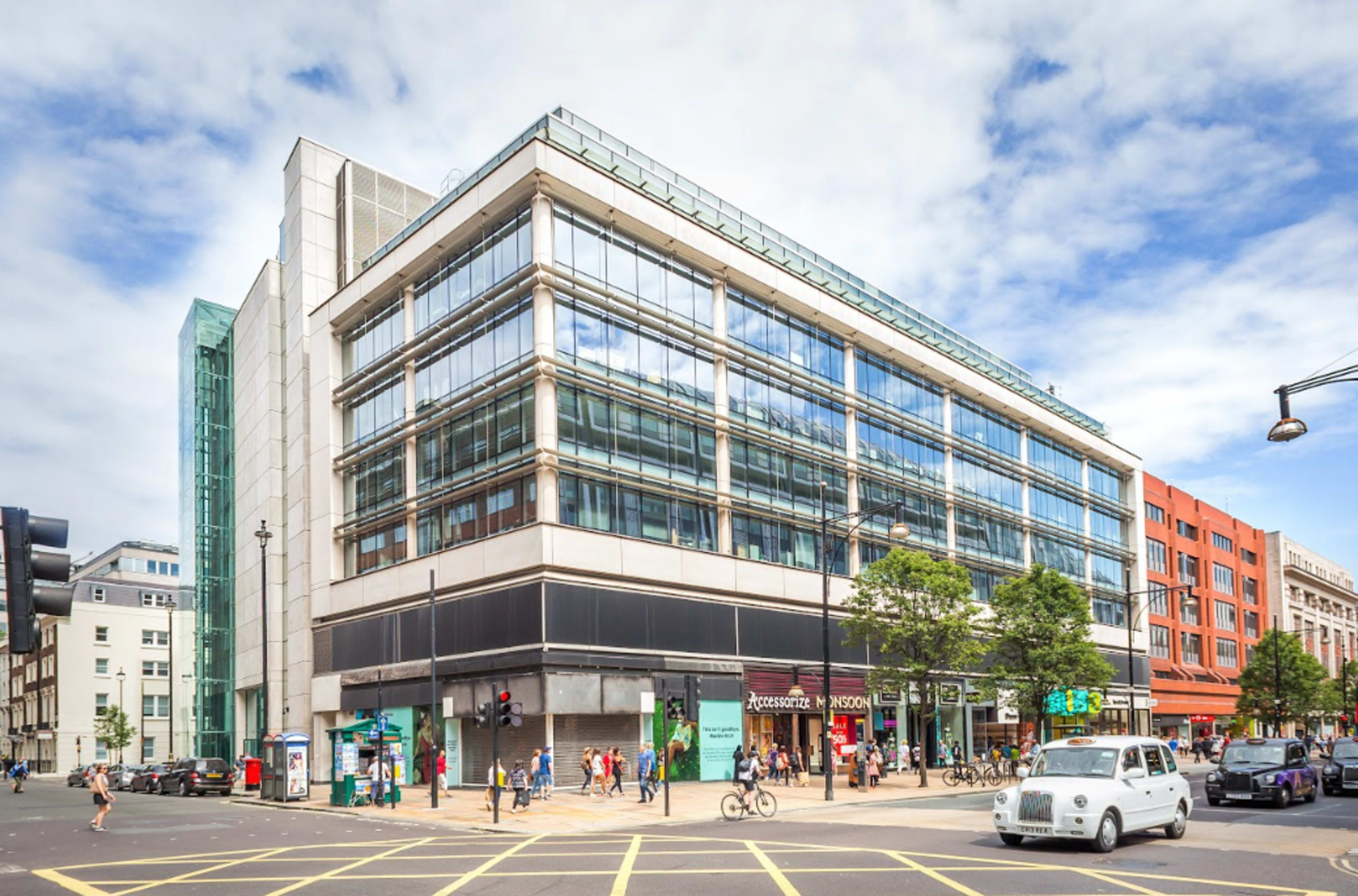 A modern glass-fronted building is situated on a bustling urban street with pedestrians, shops, and vehicles.