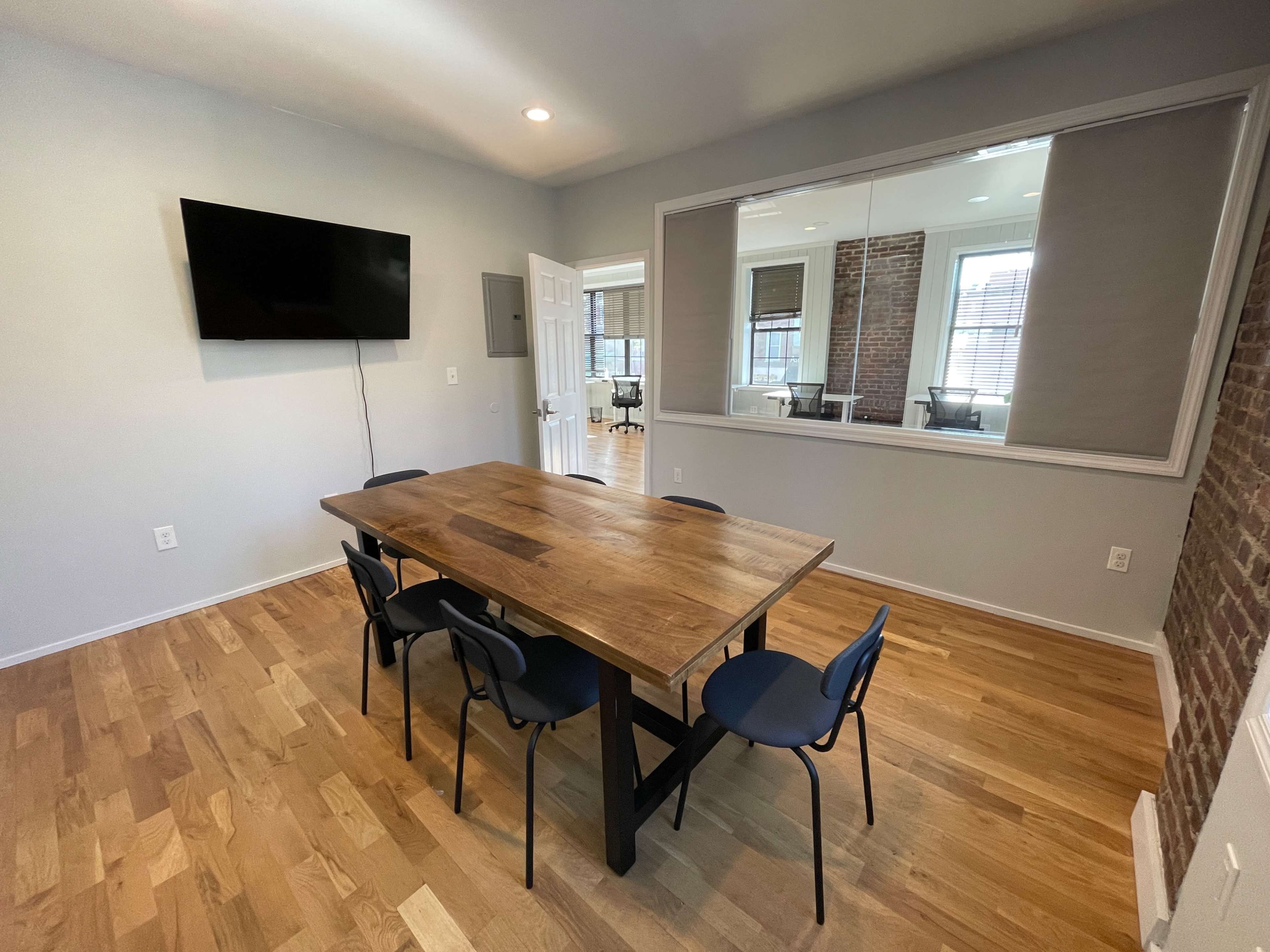 A simple, modern meeting room featuring a wooden table surrounded by chairs, with a television mounted on the wall and a large mirror reflecting part of the space.