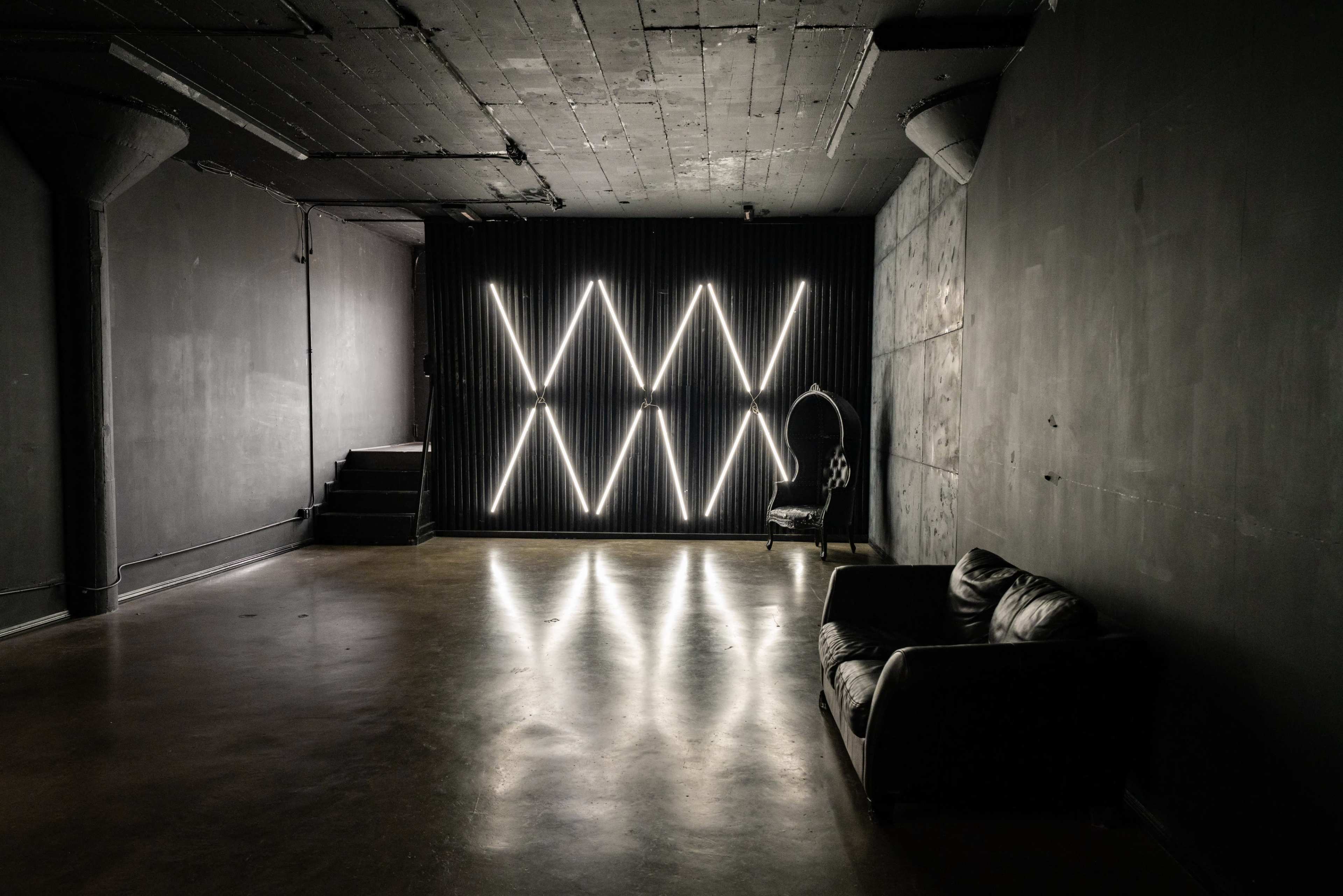 The image shows a dimly lit room with a black wall featuring geometric light patterns and a black leather couch positioned on the floor.