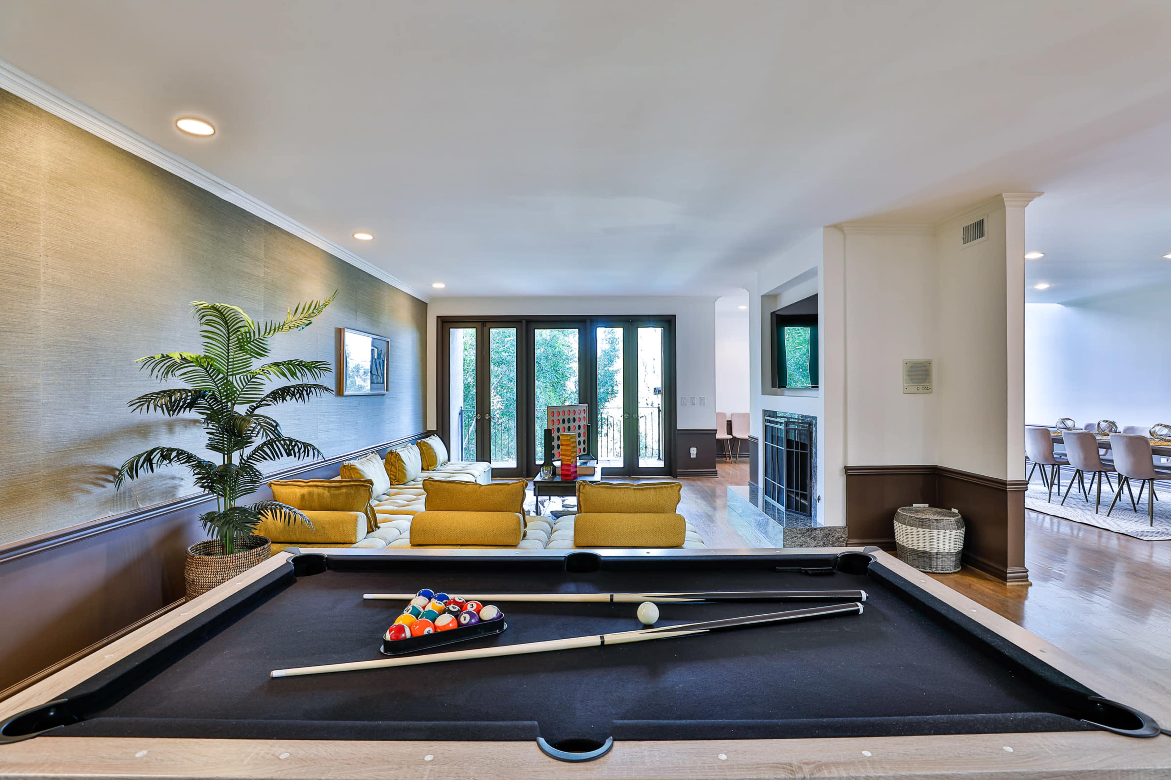 A pool table with cues and balls is positioned in the foreground of a spacious living area featuring a seating space and large windows.