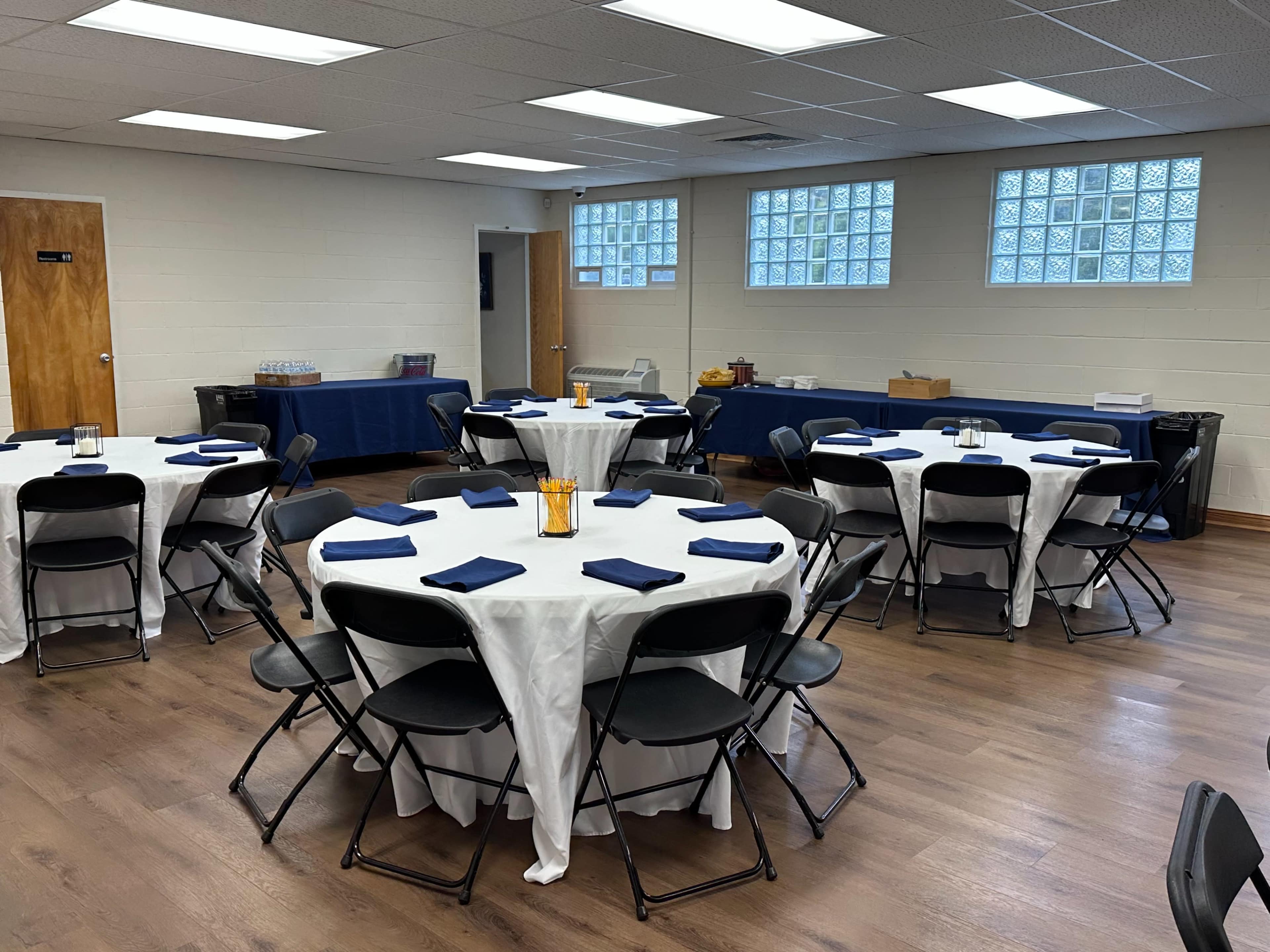 A well-lit room features several round tables covered with white tablecloths and set with blue napkins, surrounded by black chairs.