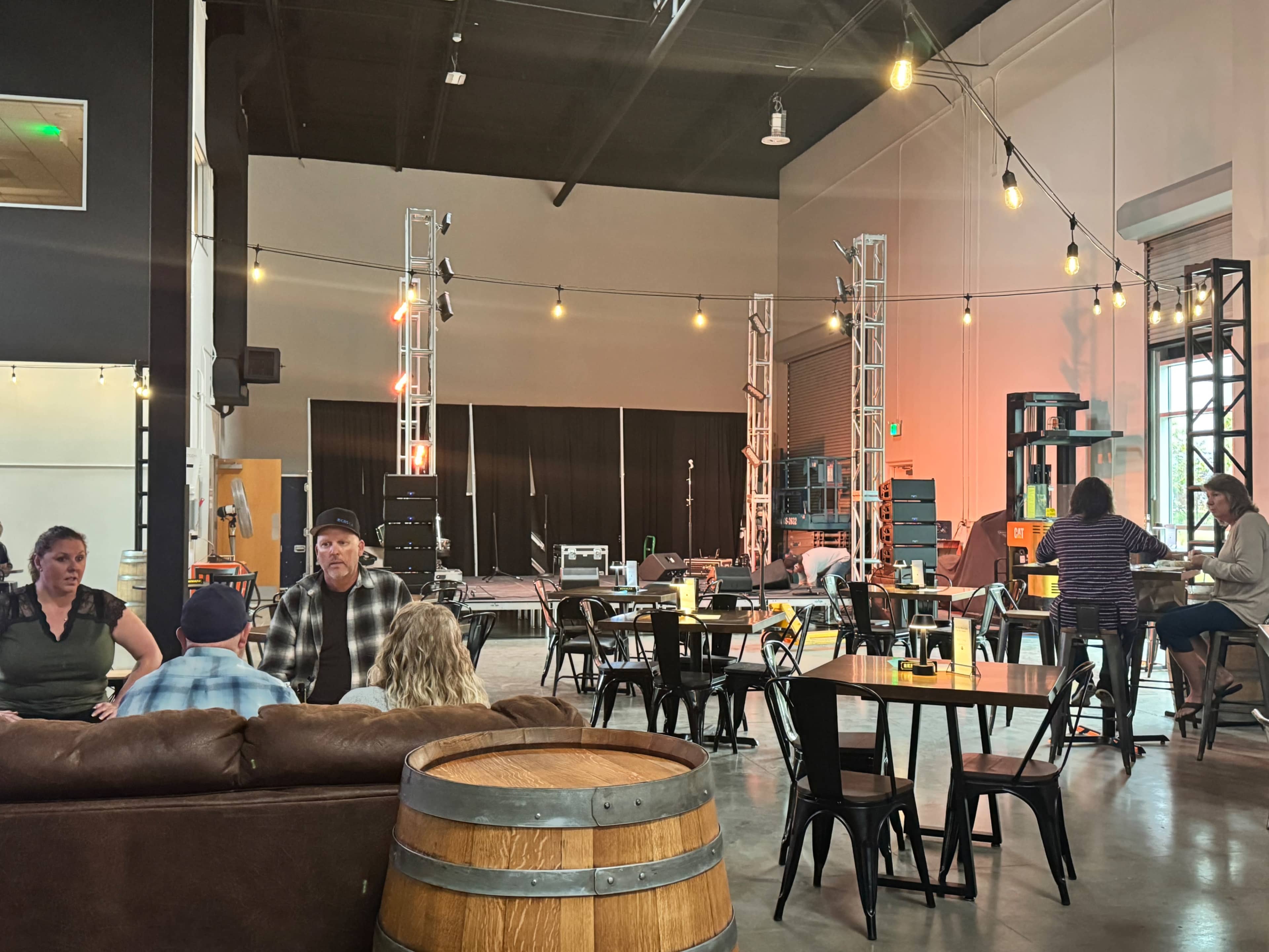 The image shows a spacious event venue with a stage, string lights, and seating areas including tables and couches, with some people gathered and engaging in conversation.