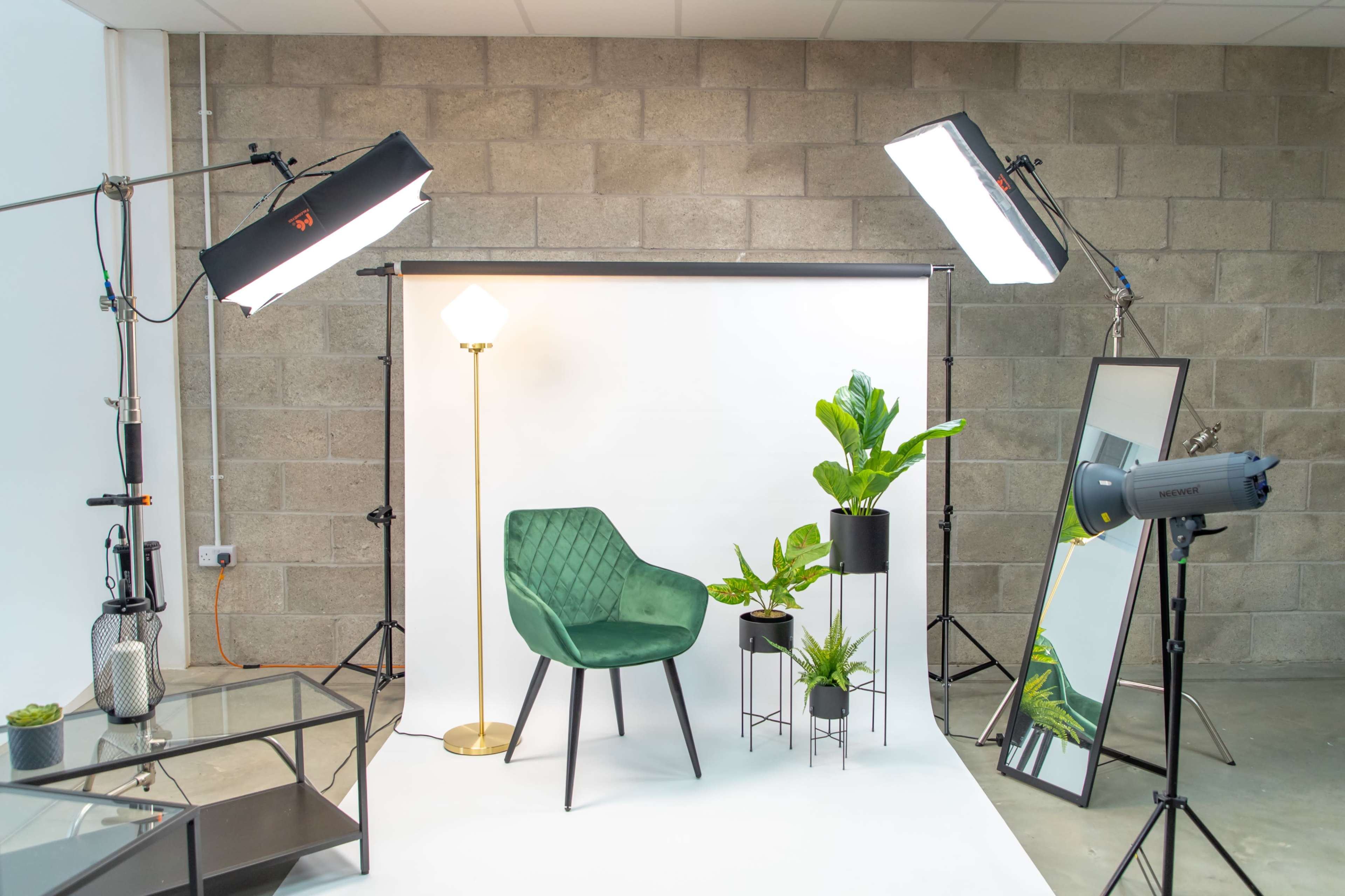A photo studio setup featuring a green chair, a floor lamp, two potted plants, and various lighting equipment against a backdrop of white and gray walls.