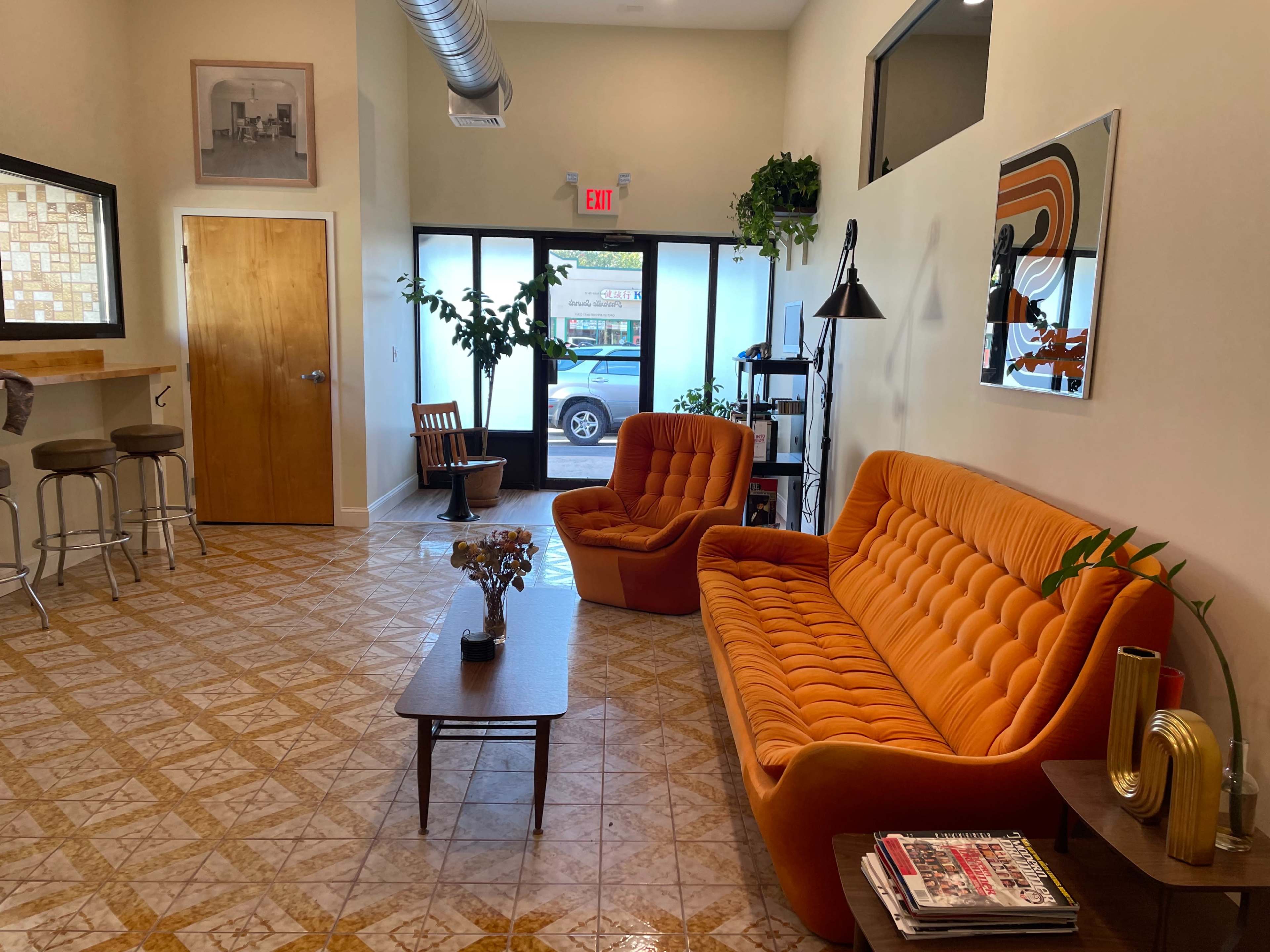 A bright, modern waiting area featuring an orange sofa, a wooden coffee table, and large windows with greenery visible outside.