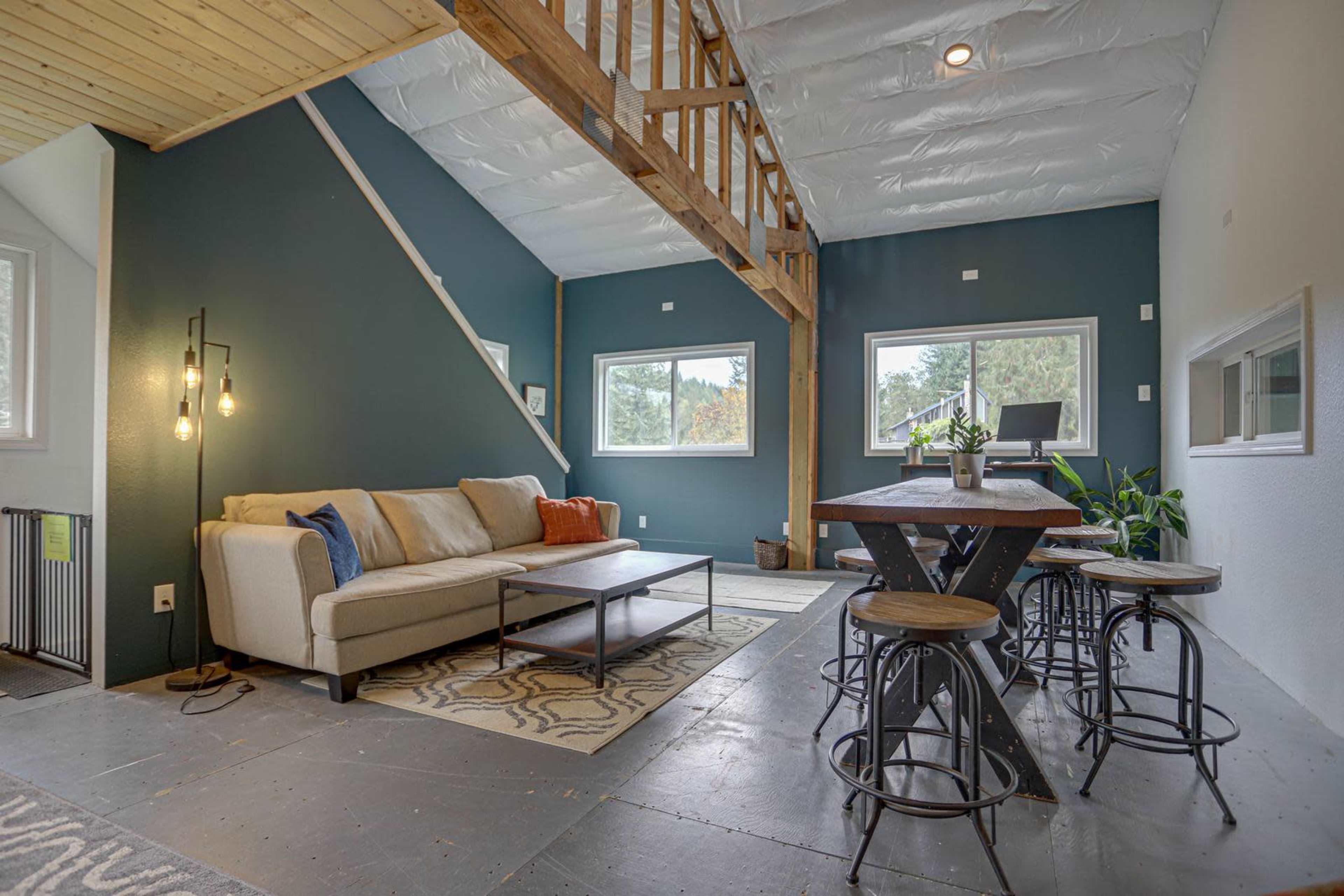 A modern living space featuring a beige sofa, a wooden table with stools, and large windows that allow natural light in.