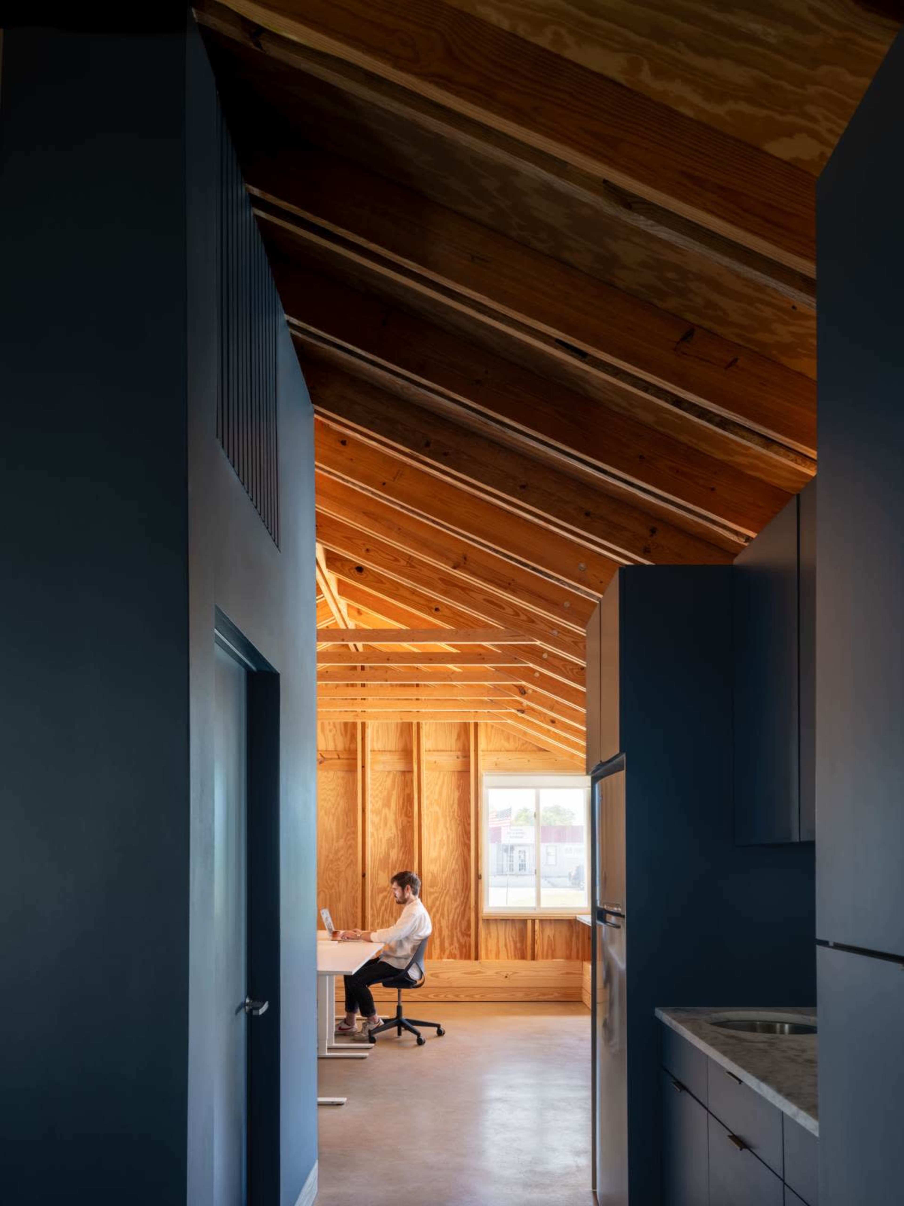 A person is sitting at a desk in a well-lit room with wooden beams and large windows, visible through a hallway with dark blue walls.