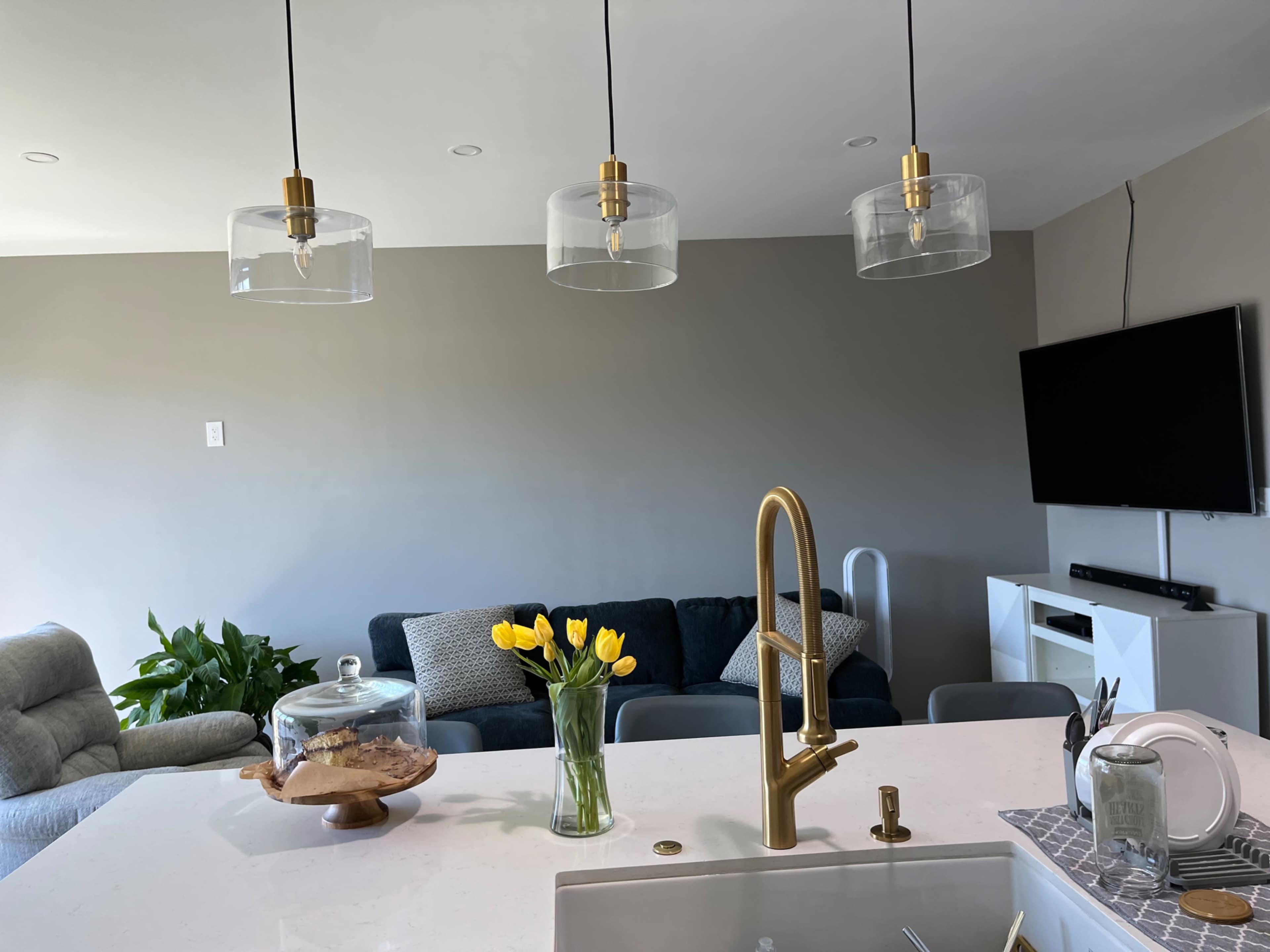 A modern living space with a kitchen island, a vase of yellow tulips, and three pendant lights hanging above, complemented by a couch and a wall-mounted TV in the background.