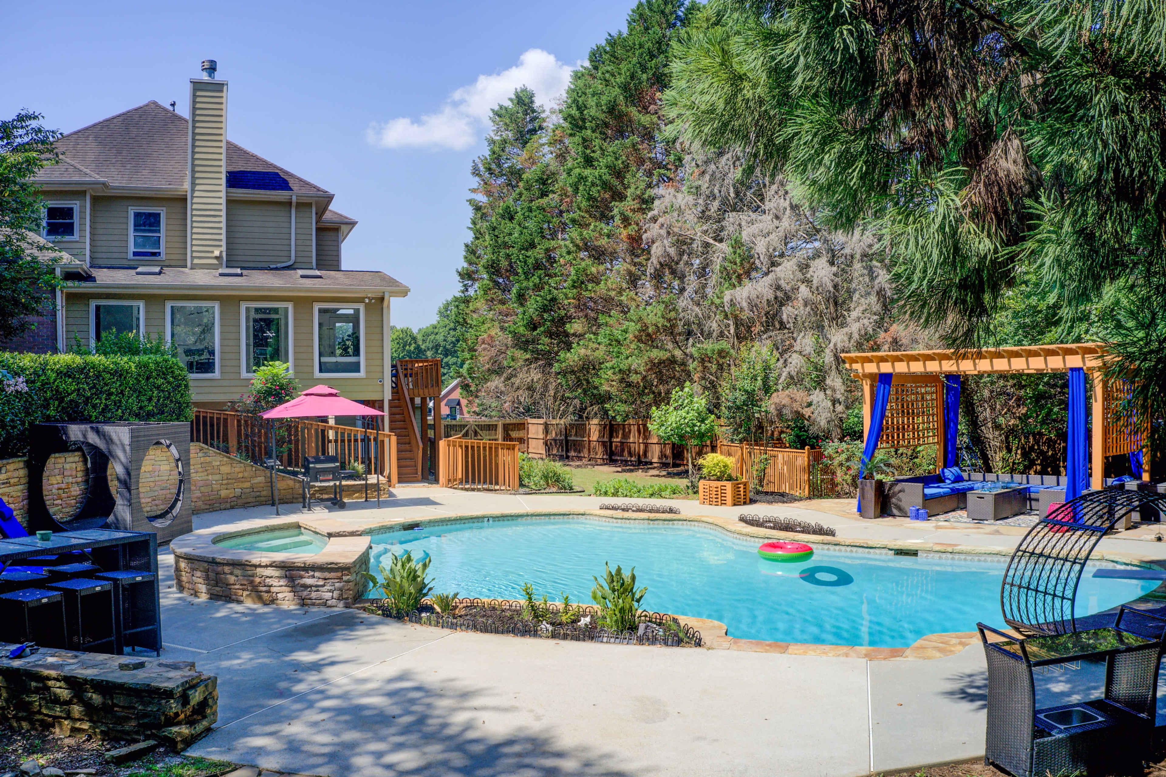 A backyard pool area features a swimming pool surrounded by landscaping, patio furniture, and a house in the background.
