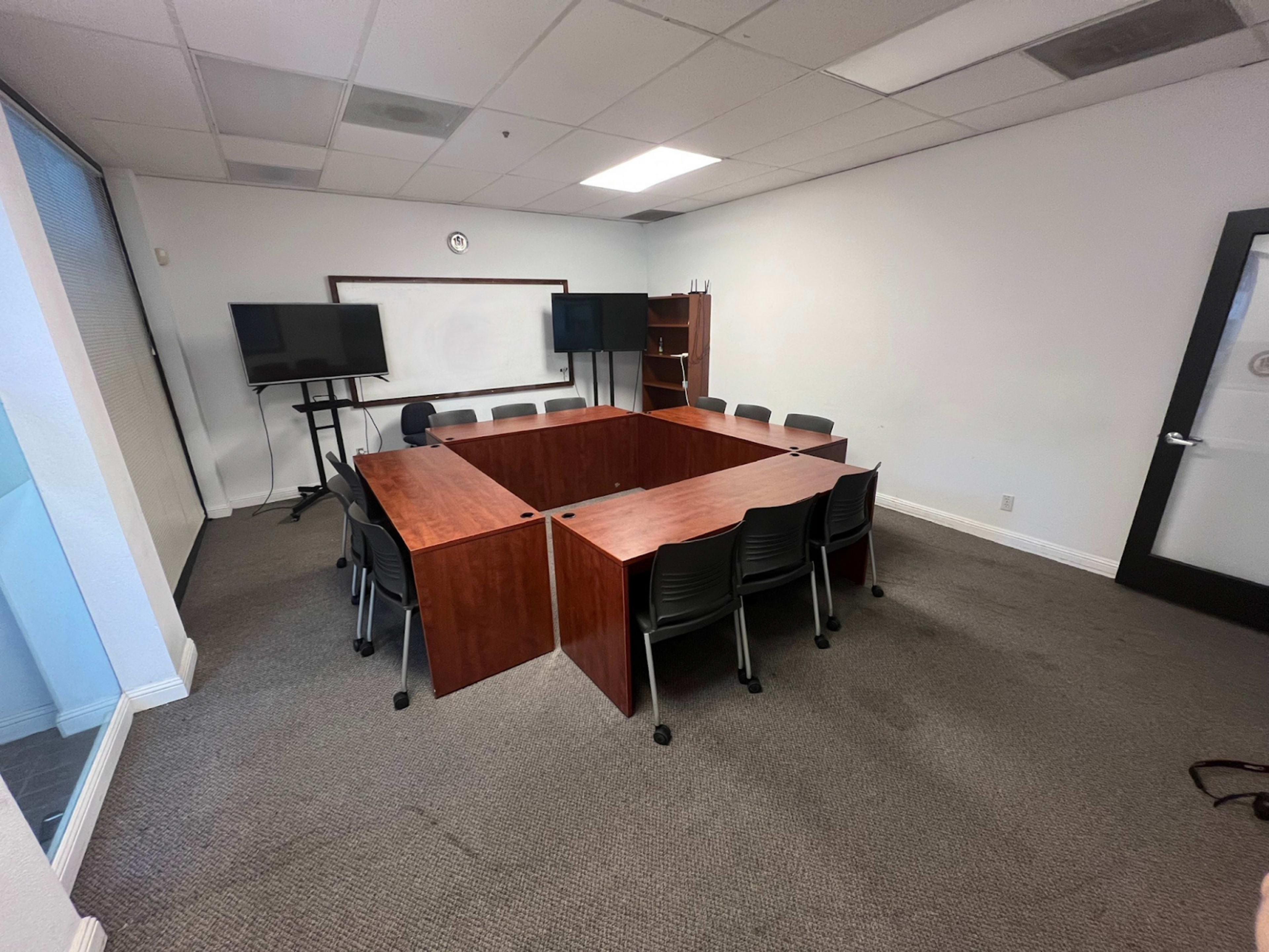 A conference room features a large wooden table surrounded by black chairs, with two screens and a bookshelf against the wall.