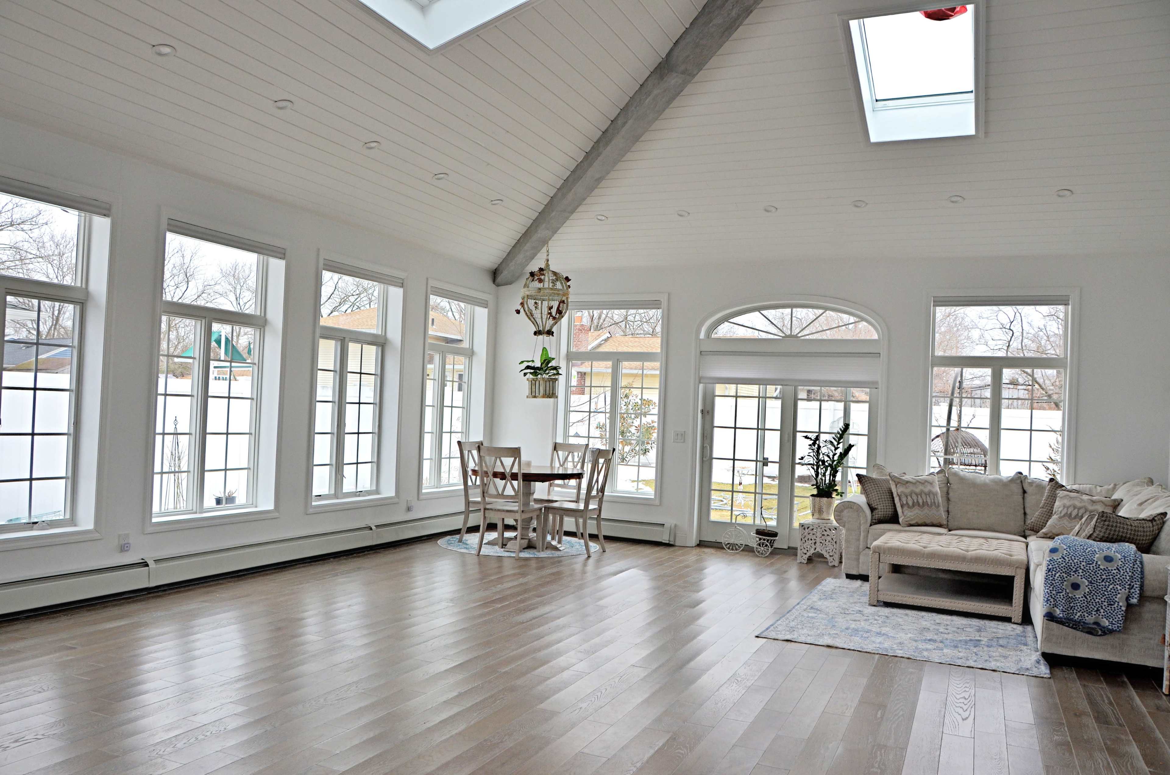 A spacious sunroom with large windows, a dining table, and a comfortable seating area, all set on wooden flooring.