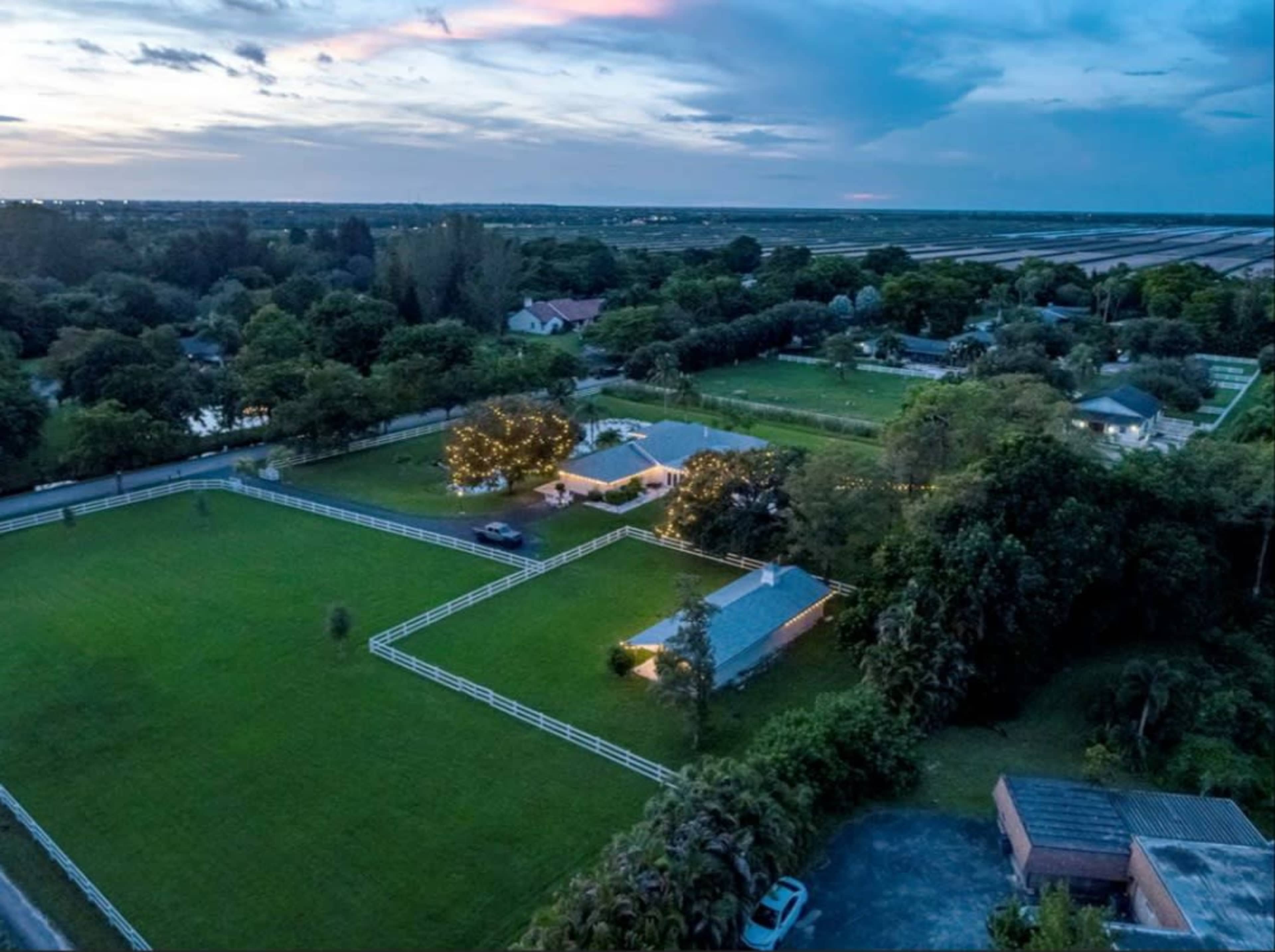 The aerial view shows a rural landscape with a house and outbuildings surrounded by lush greenery and a fenced yard under a twilight sky.