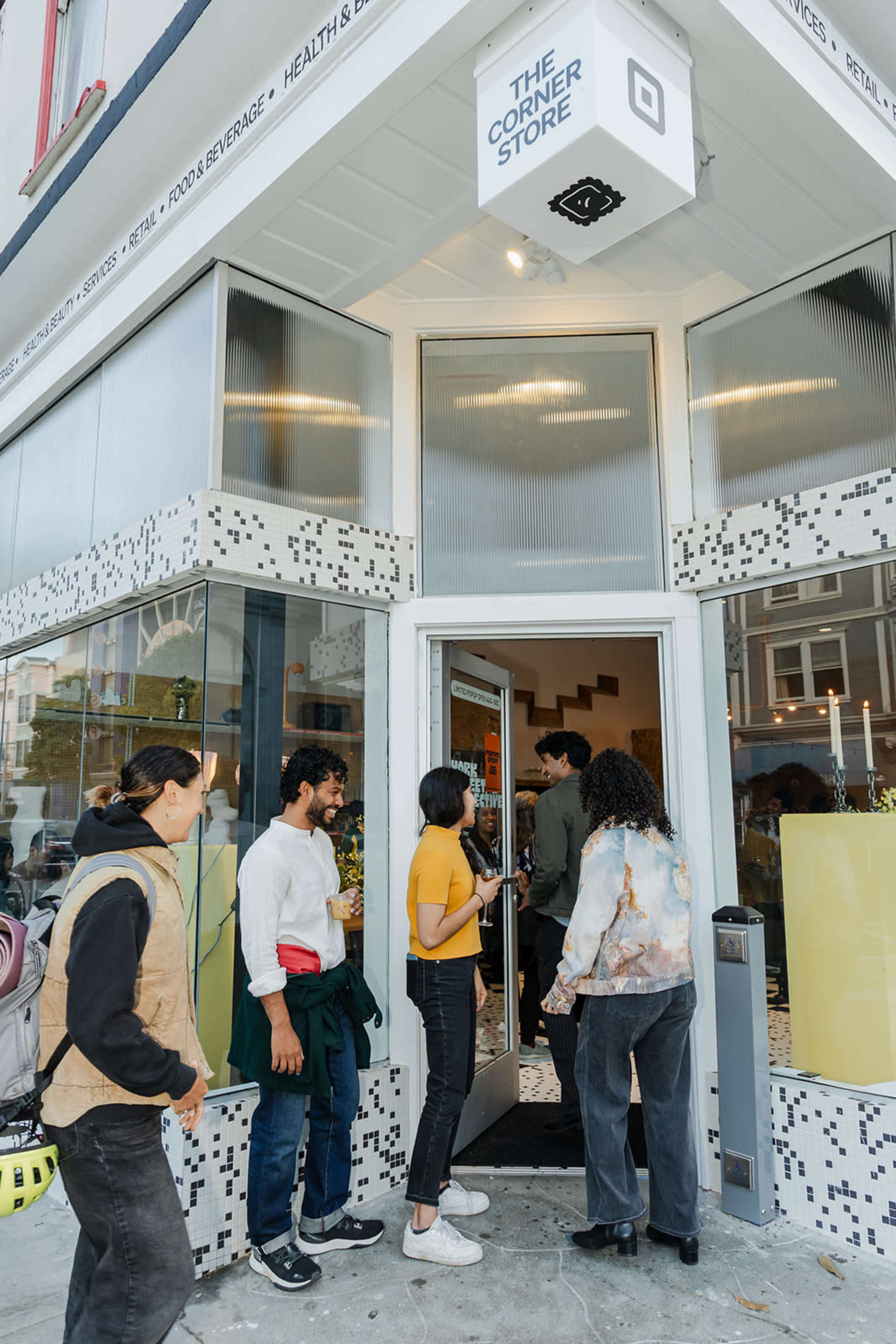 A group of people stands in line at the entrance of a shop called "The Corner Store."