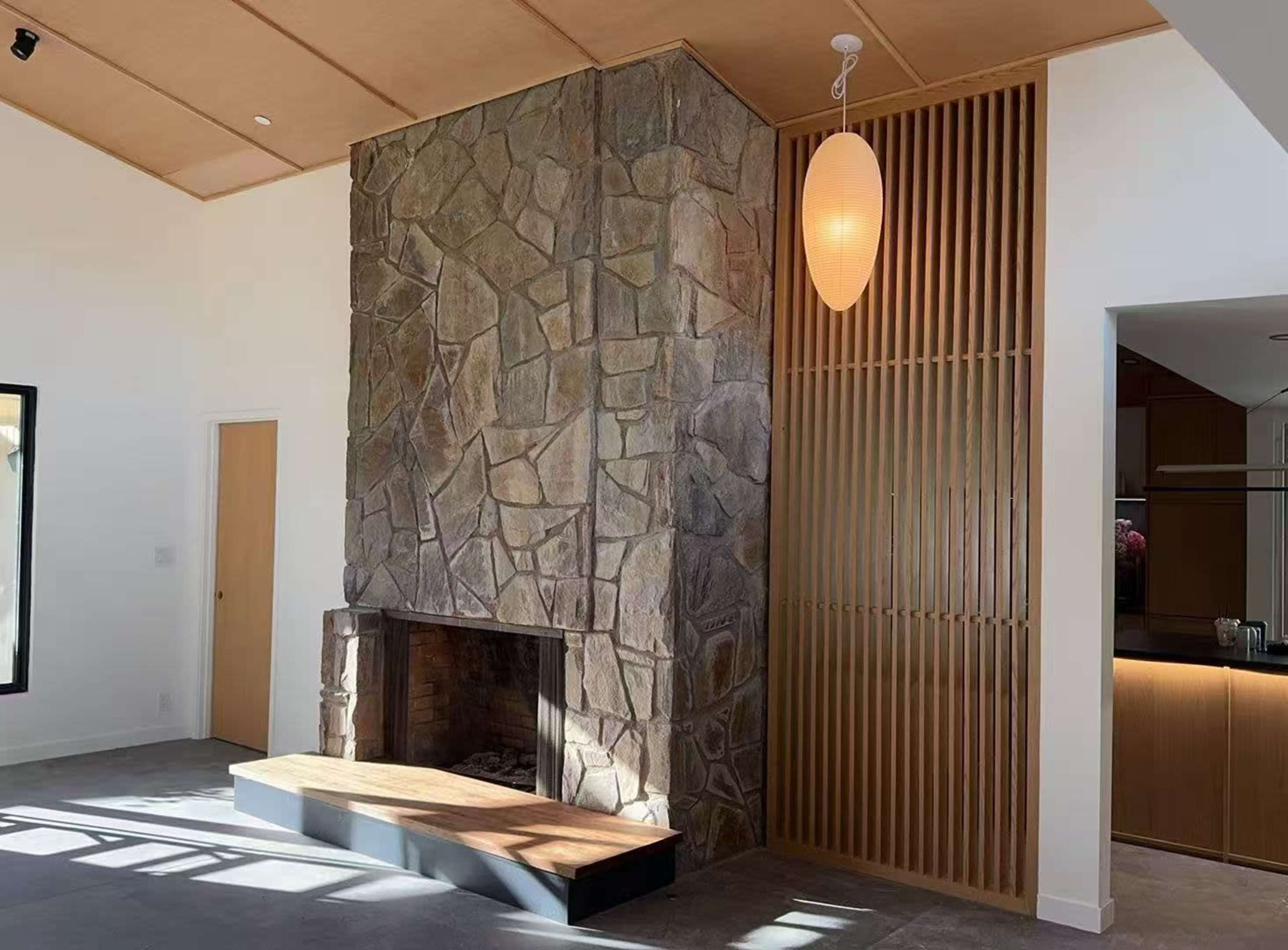The image shows a modern interior featuring a large stone fireplace against a wall, paired with wooden paneling and a pendant light.