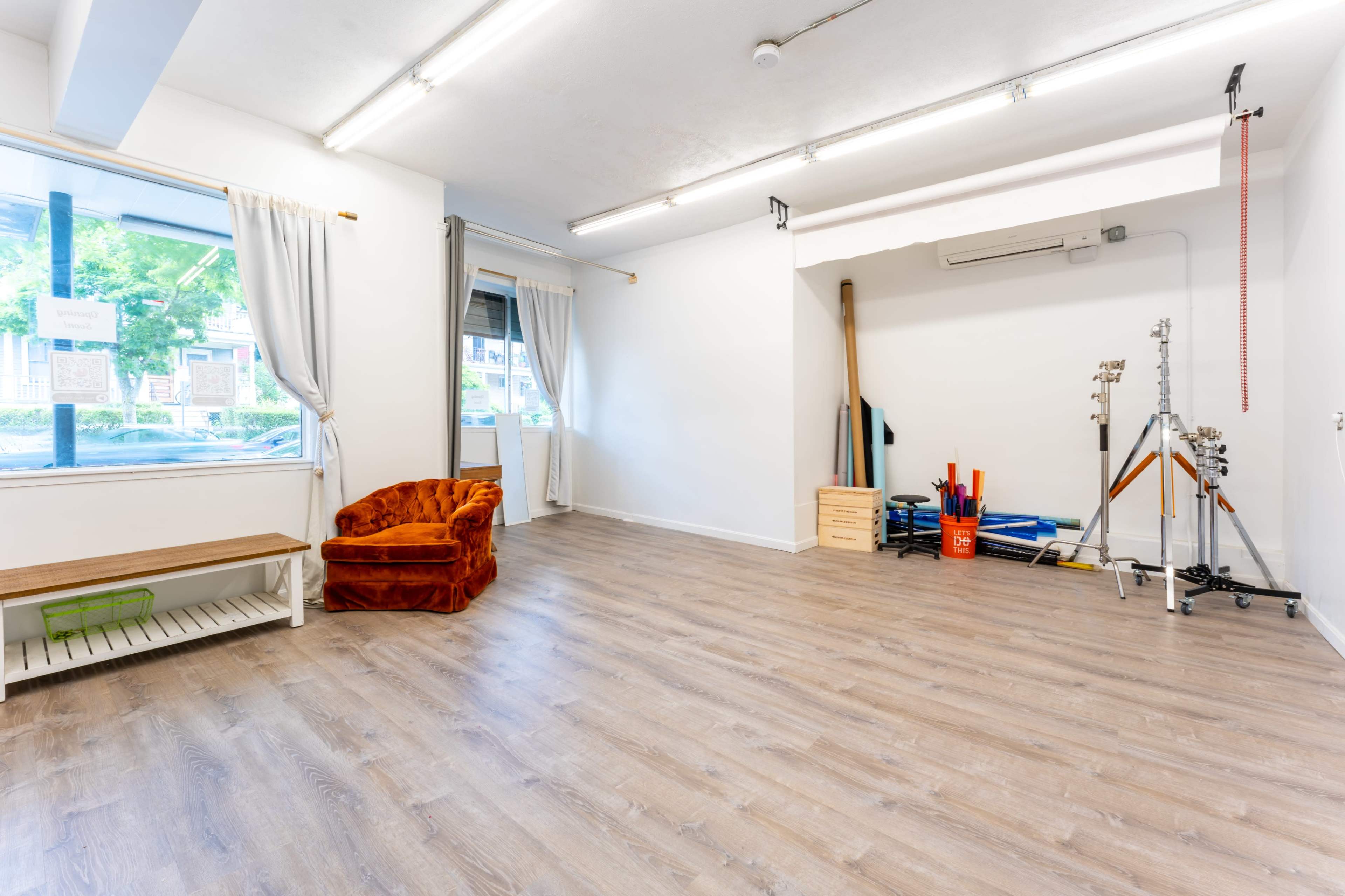 A bright, sparsely furnished room with wooden flooring, featuring a velvet orange chair, a window with curtains, and various exercise equipment in the corner.