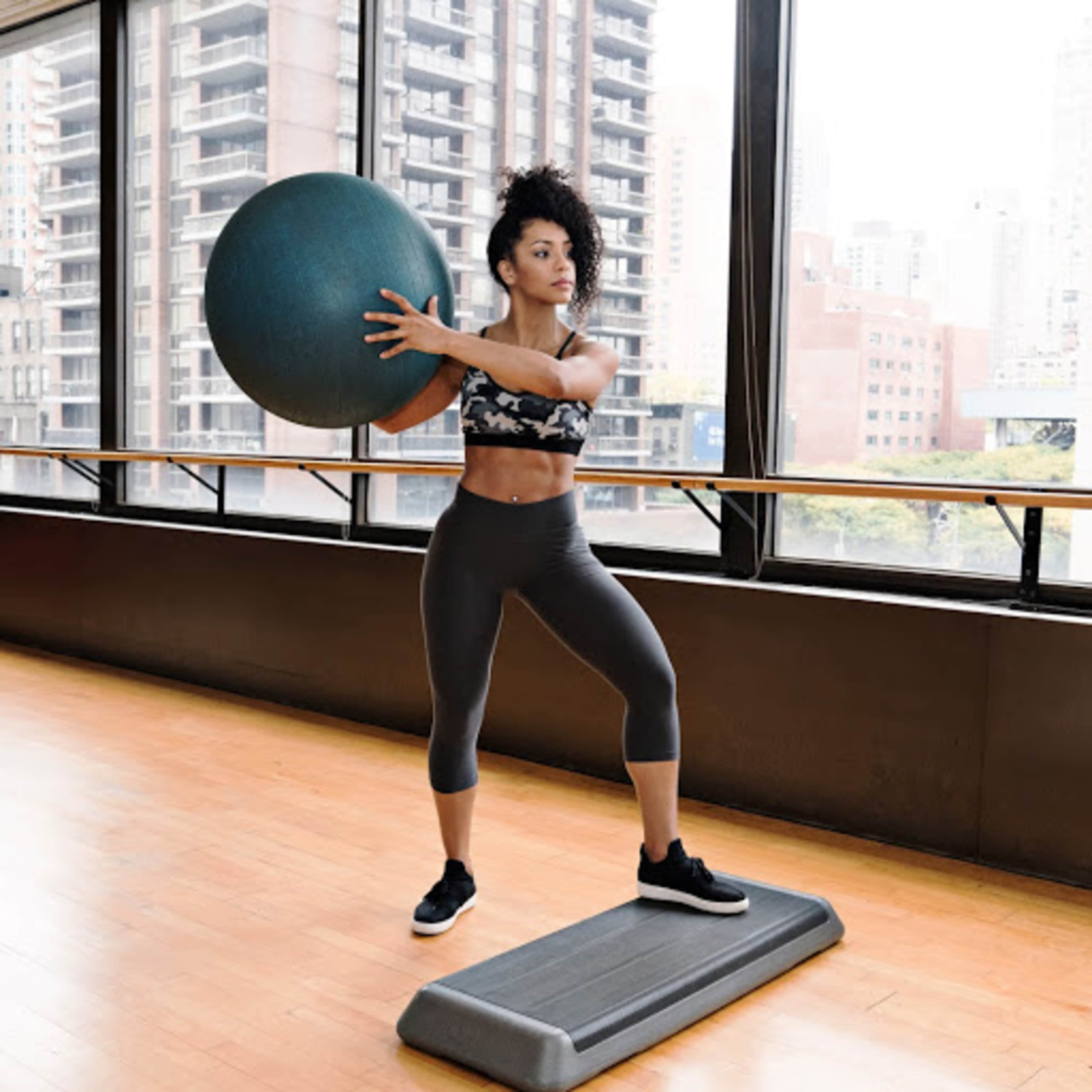 A woman is performing an exercise with a stability ball while standing on a fitness step in a brightly lit gym.