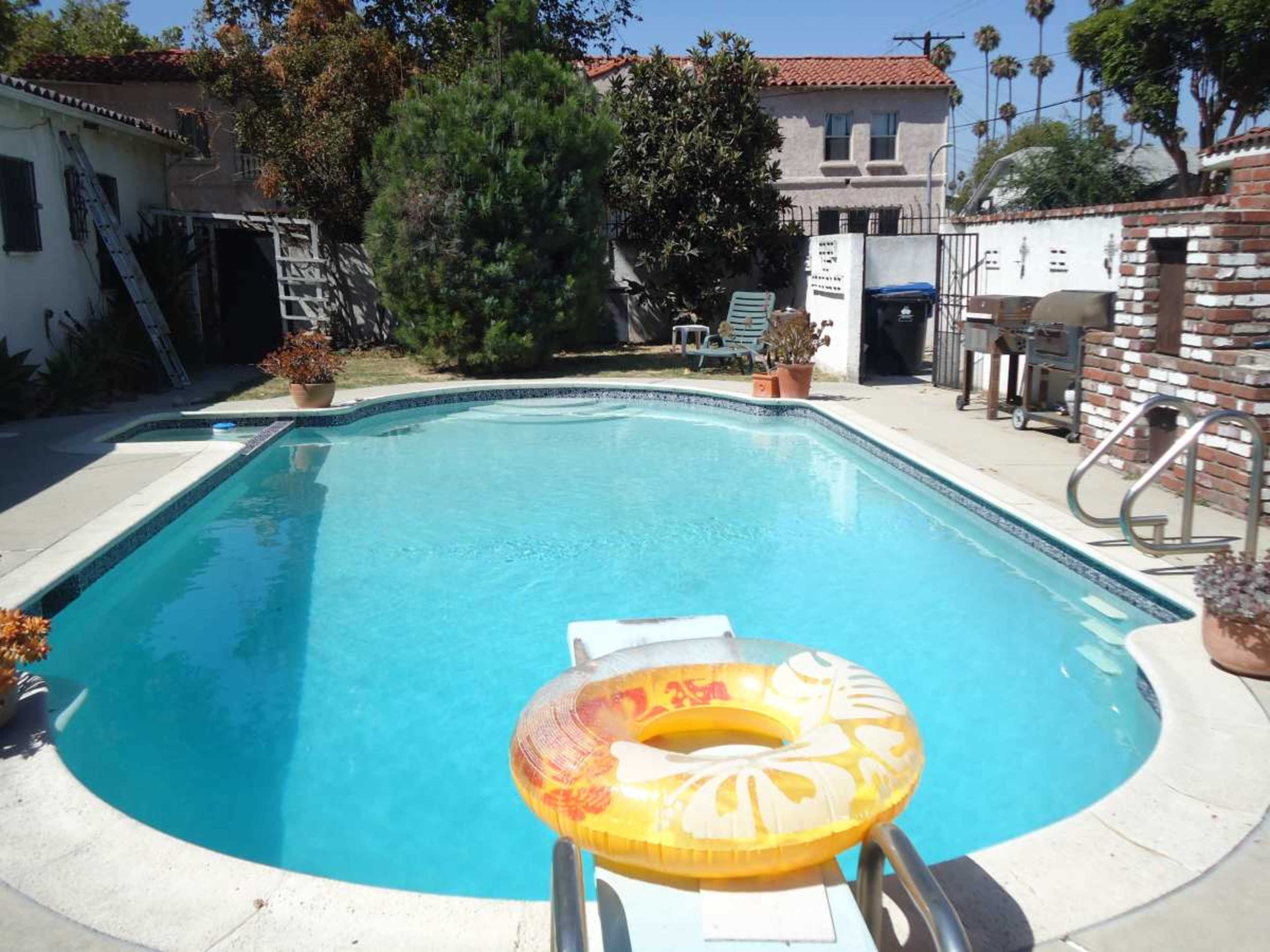 The image shows a clear swimming pool with a yellow inflatable ring on the edge, surrounded by a patio area that features potted plants and outdoor furniture.
