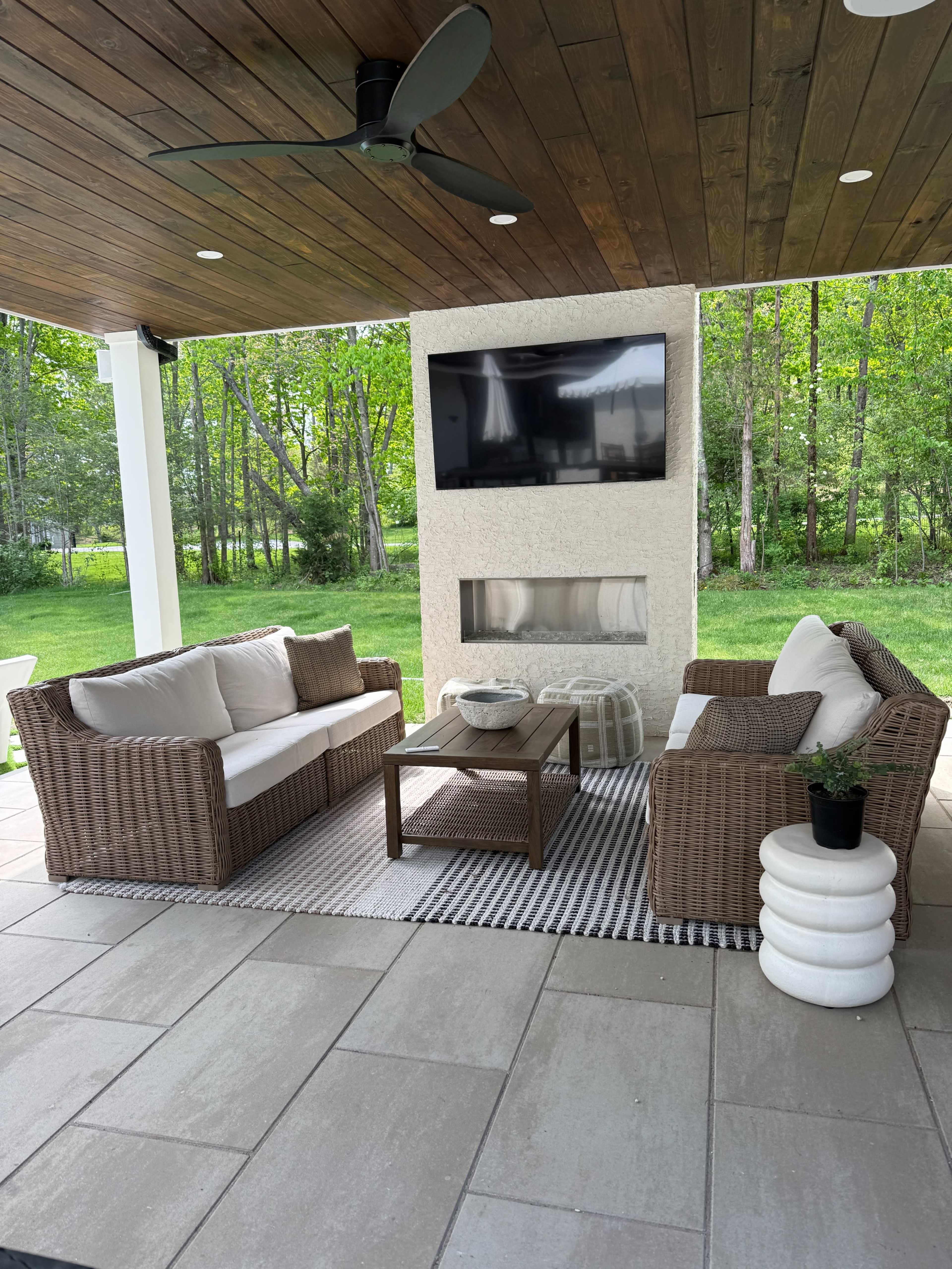 The image shows an outdoor seating area with two wicker sofas, a coffee table, and a mounted TV, surrounded by lush greenery.