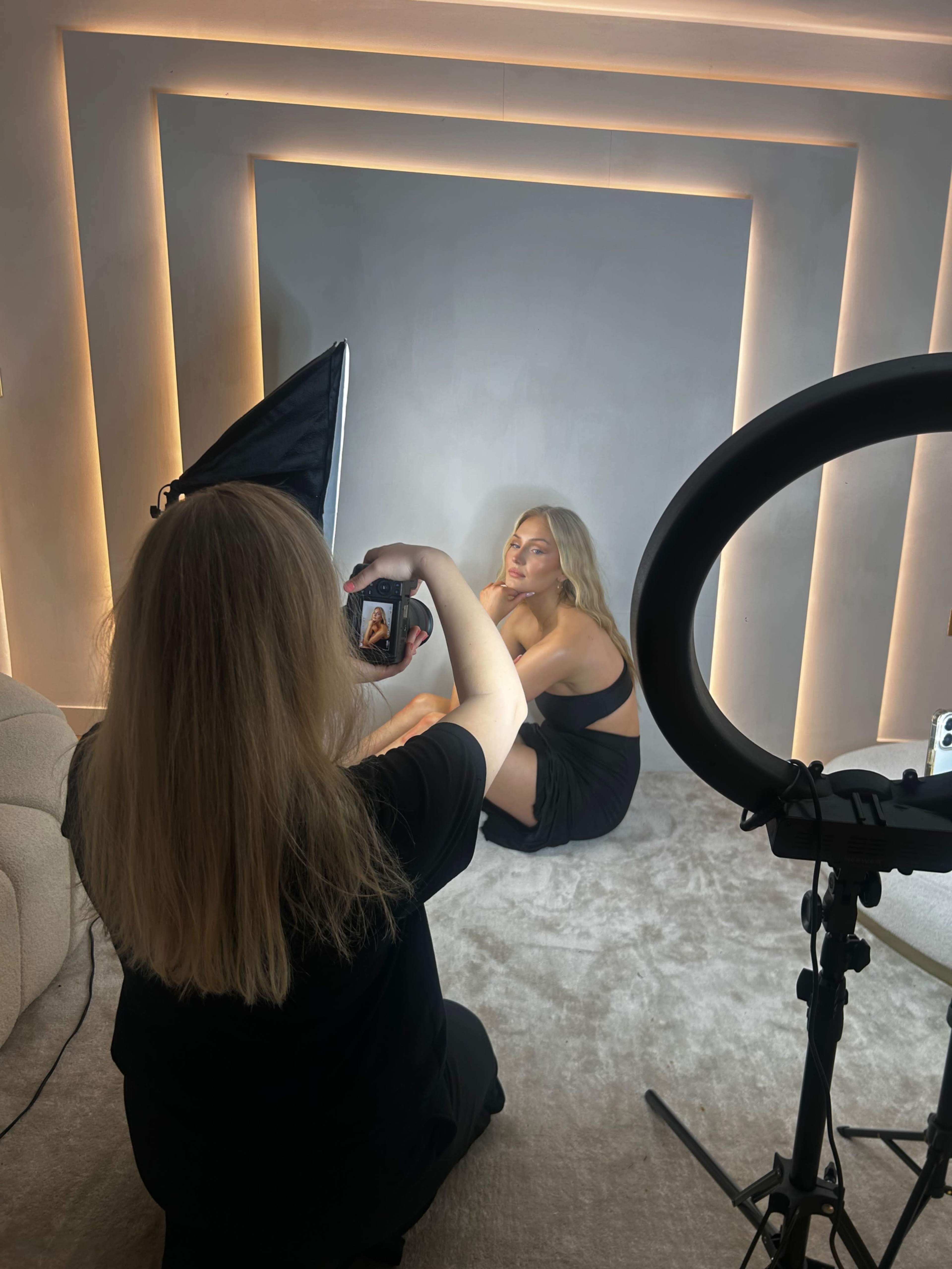 A woman is photographing another woman posing against a softly lit backdrop in a studio setting.