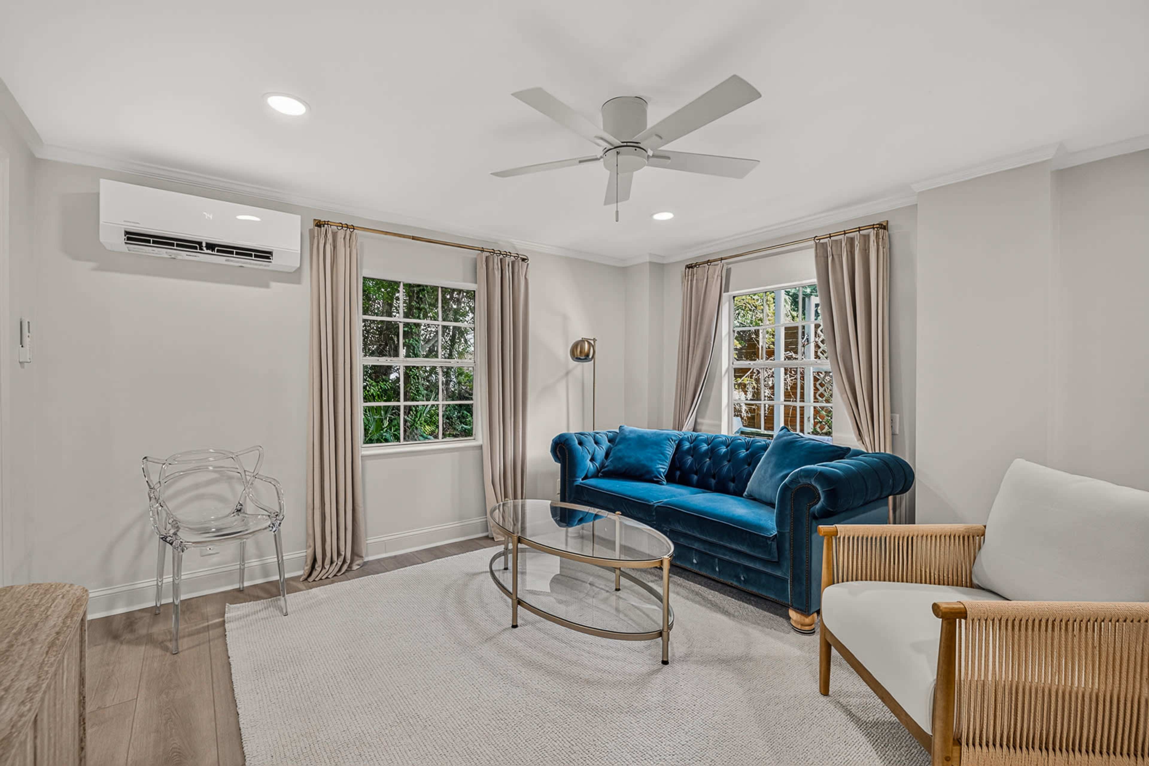 The image showcases a modern living room featuring a blue velvet sofa, two windows, a glass coffee table, and a combination of light-colored curtains and seating.