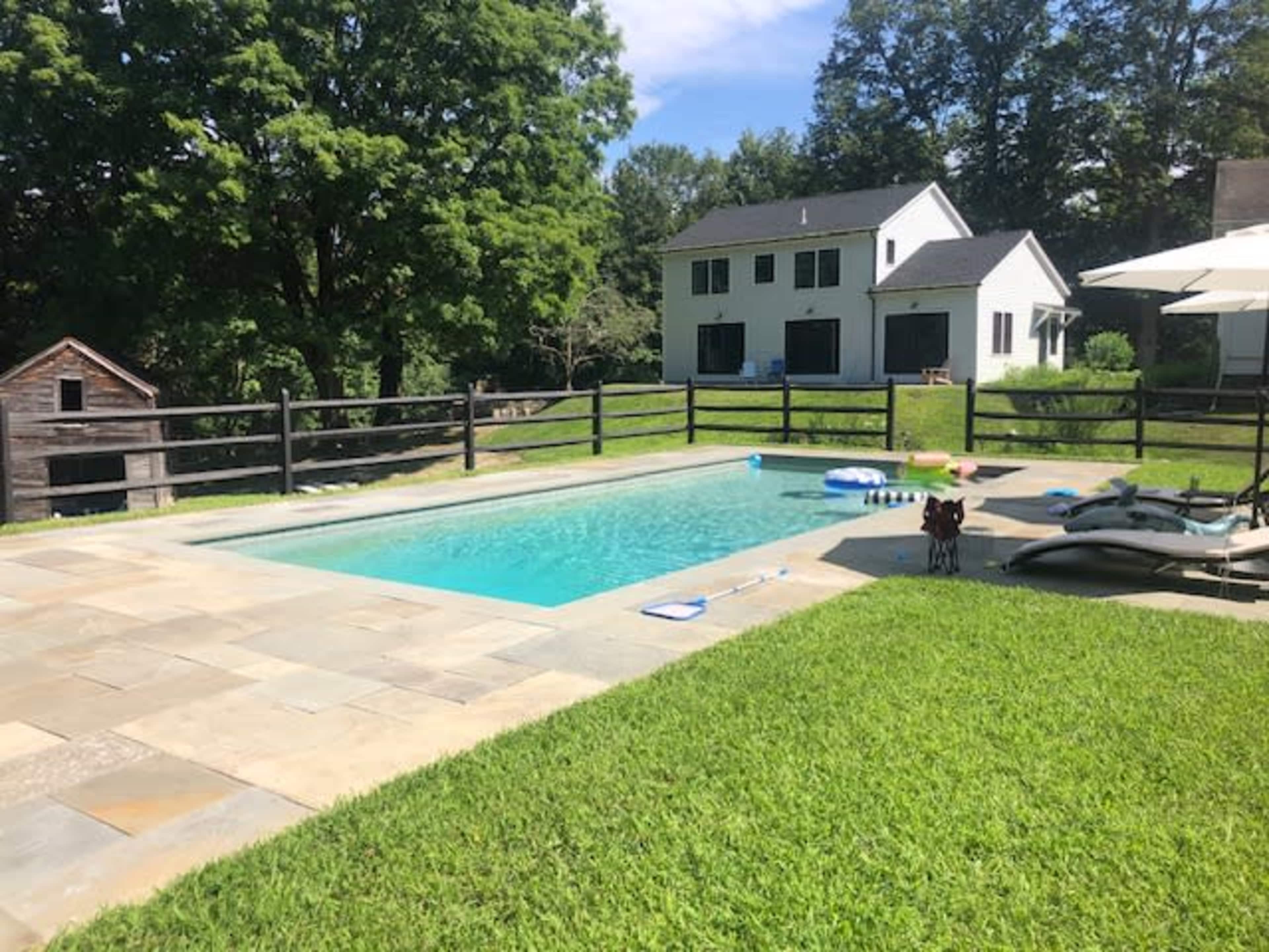 The image shows a rectangular swimming pool surrounded by a stone patio and green grass, with a house in the background and a small shed to the left.