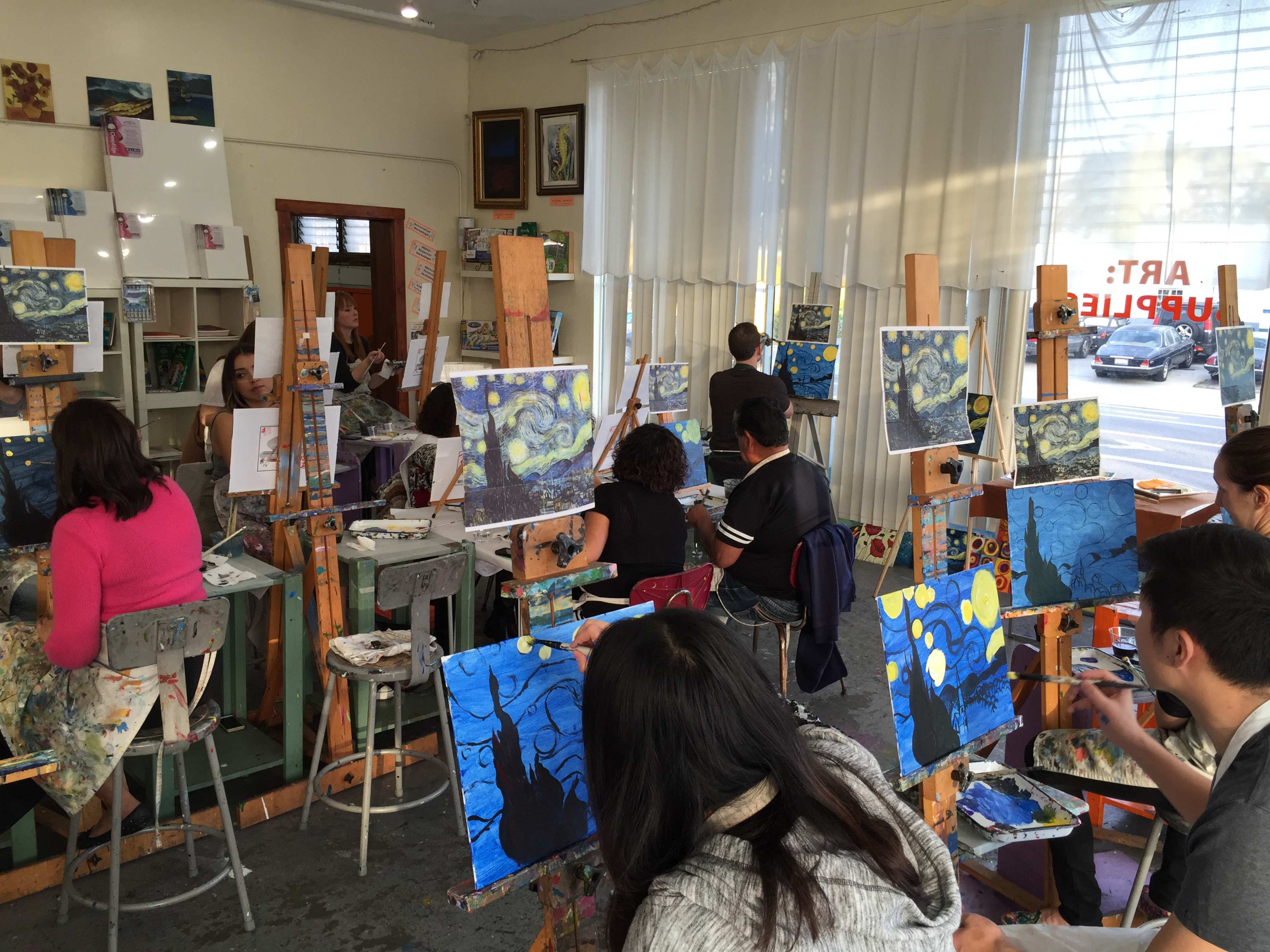 A group of people is painting at easels in a bright art studio, working on their interpretations of a starry night scene.