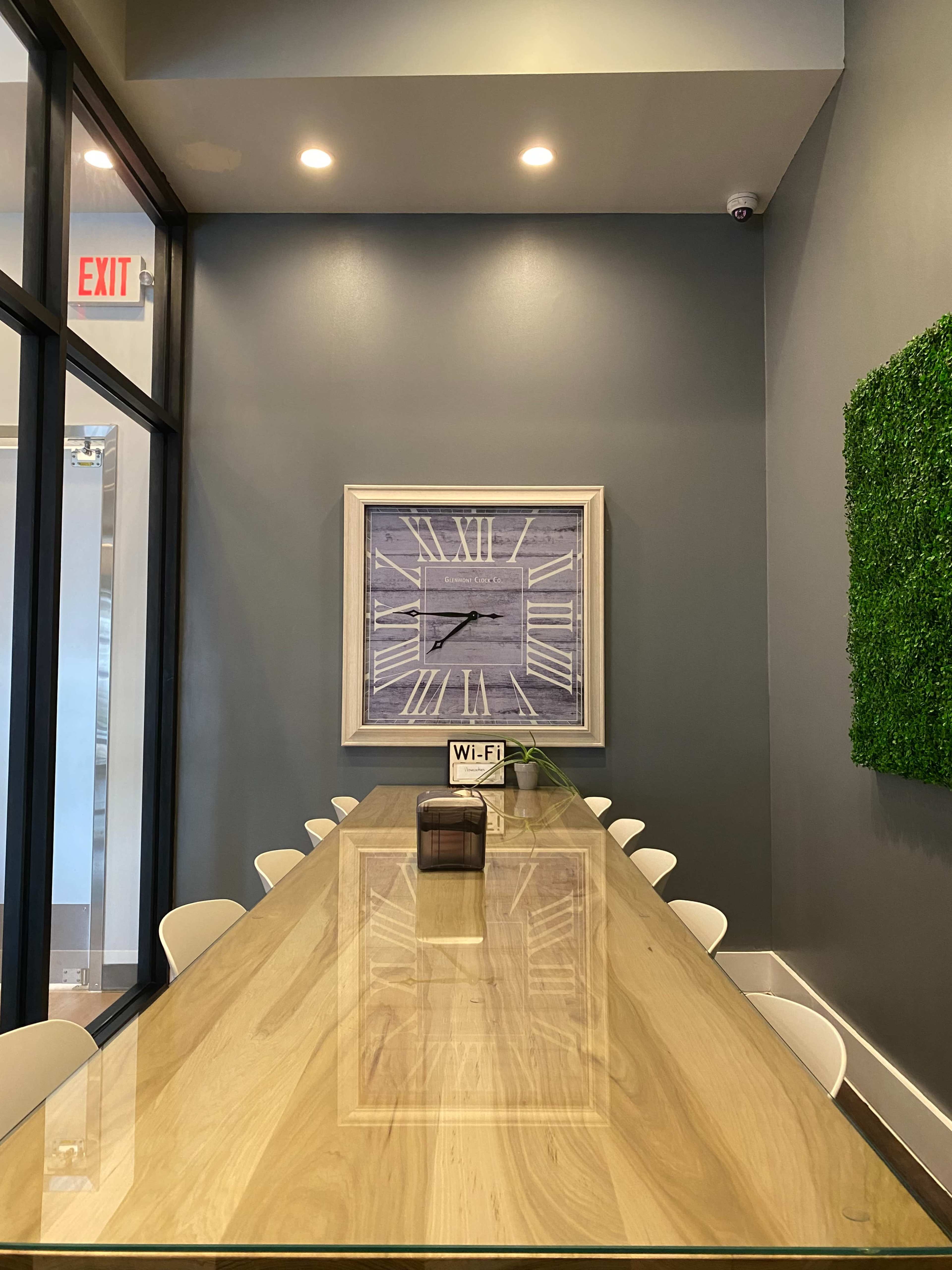 A long wooden table with white chairs is positioned in a well-lit room featuring a large wall clock and a green wall accent.