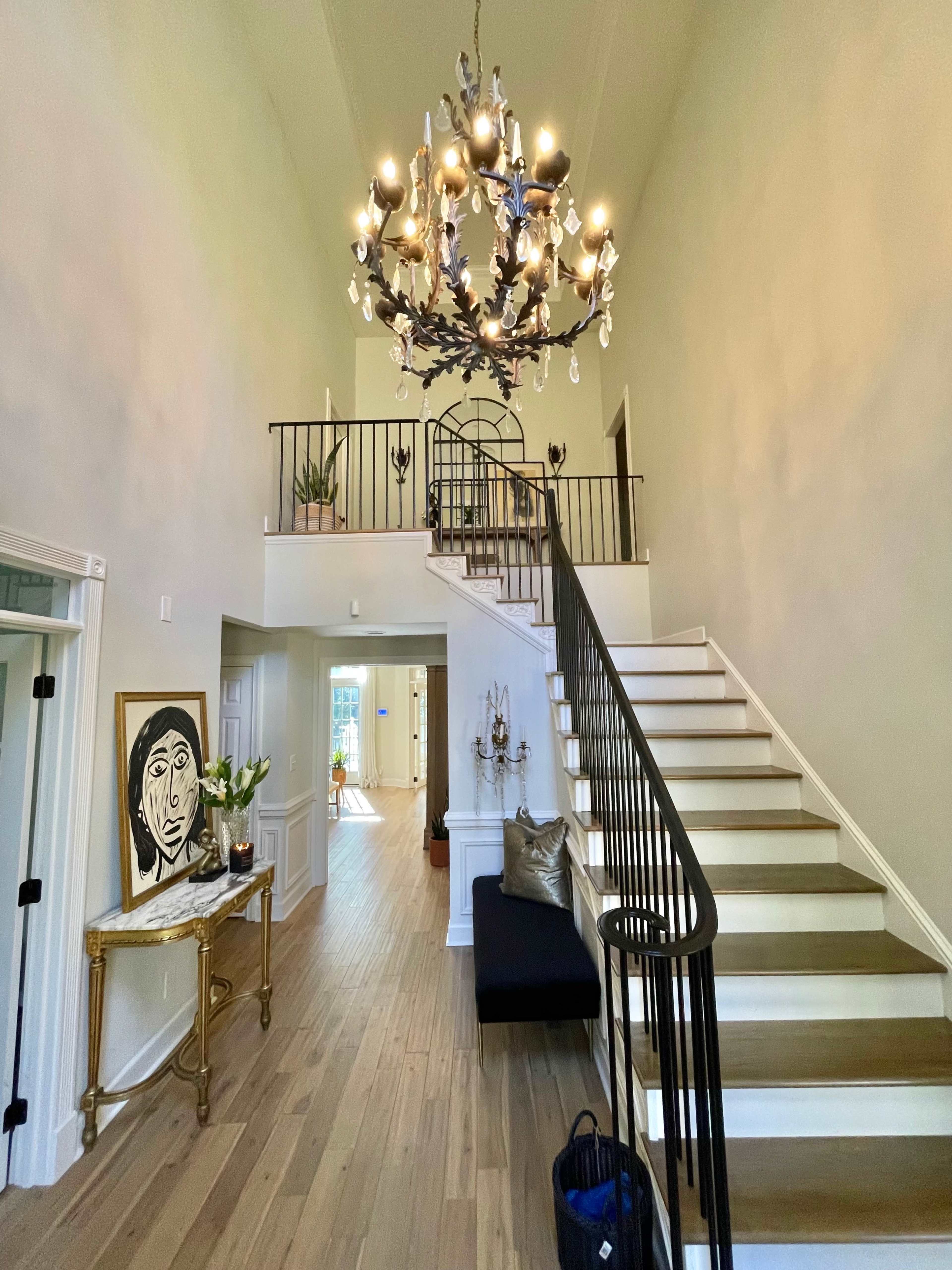 A spacious foyer features a curved staircase, a large chandelier, and a decorative table with artwork.