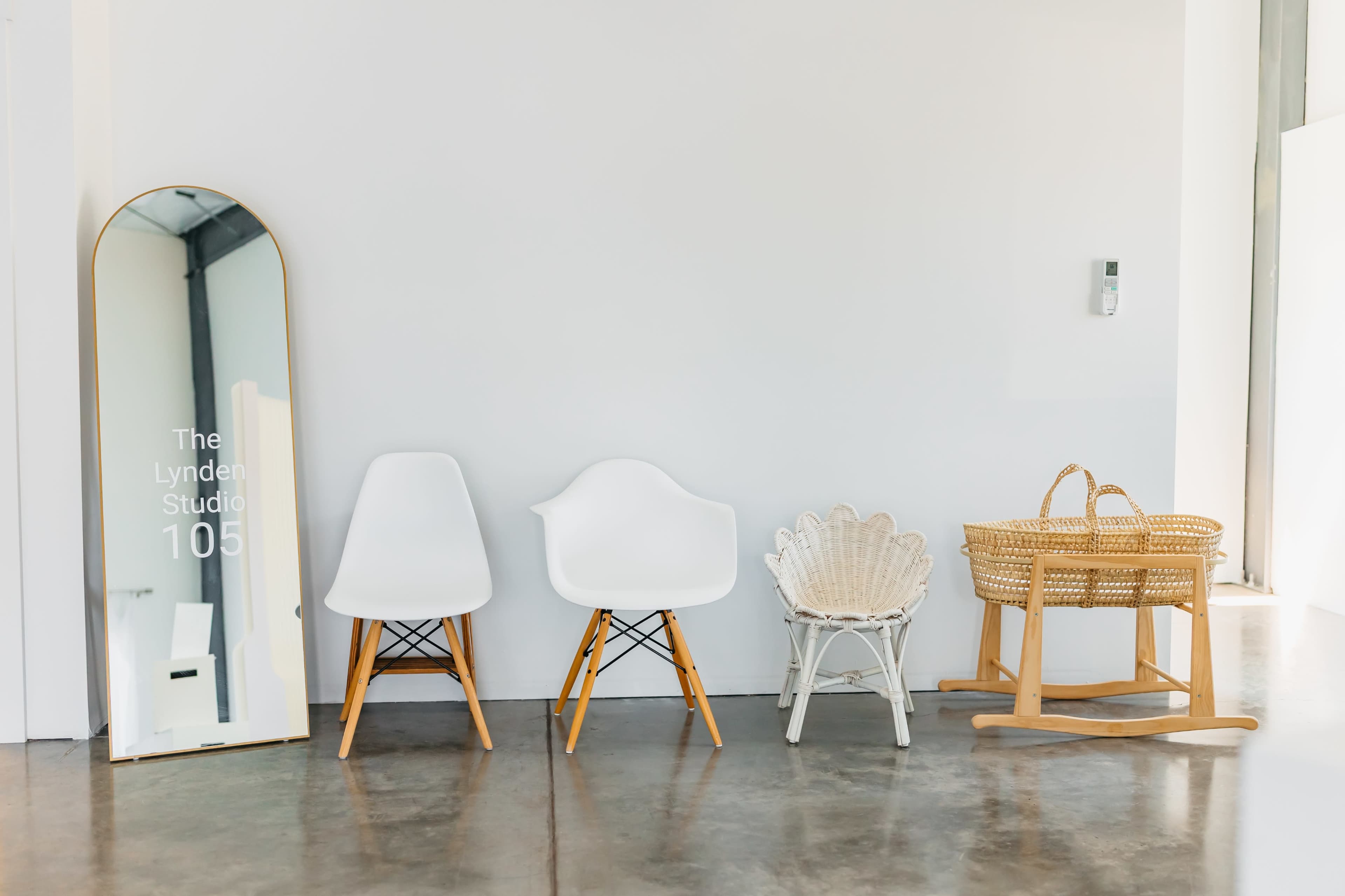 The image shows a minimalist interior with a large mirror and four different chairs, including two white plastic chairs, a woven chair, and a rocking chair made of wicker.
